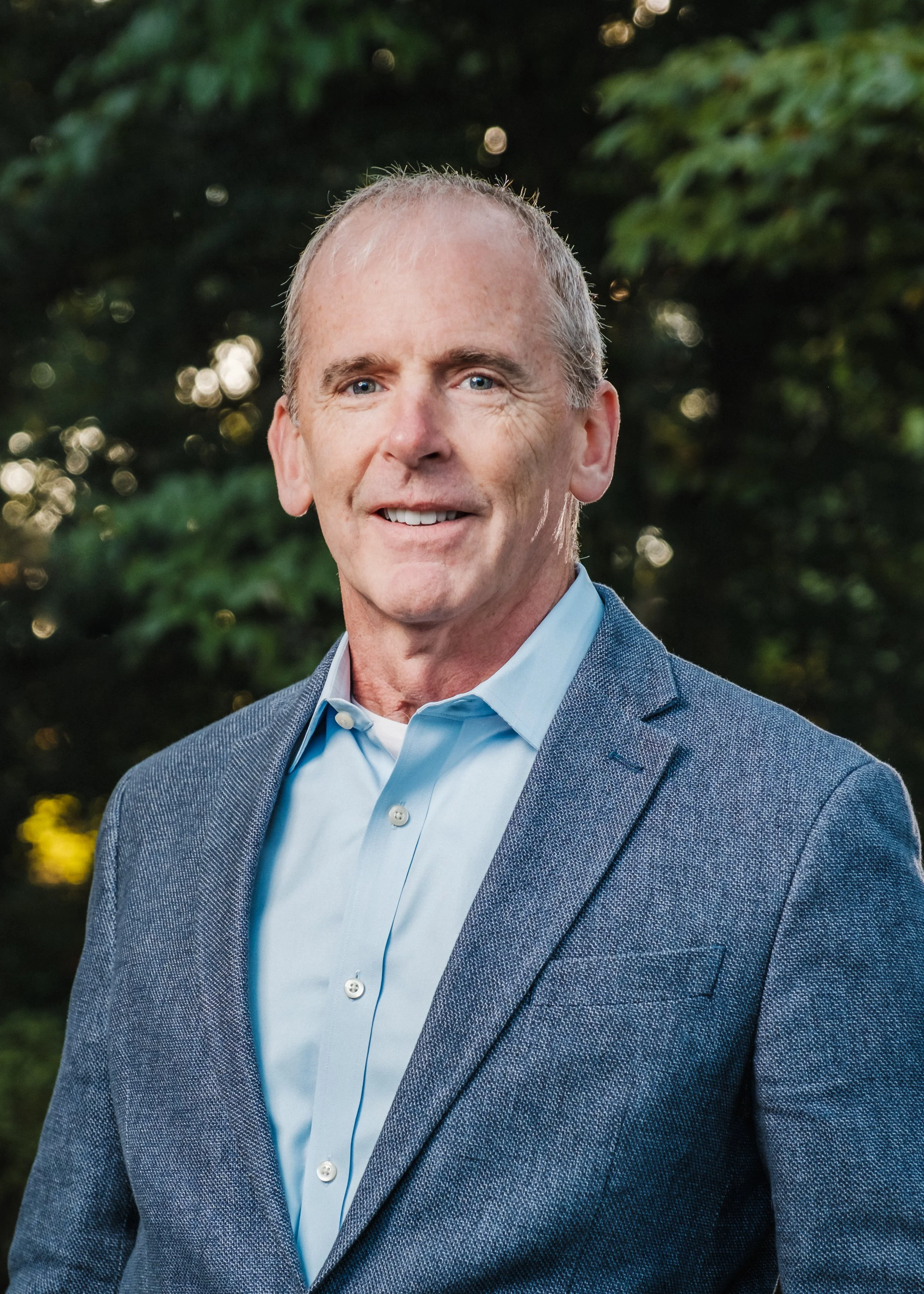 Portrait of a middle-aged man with short light hair, wearing a blue blazer and light blue collared shirt, standing outdoors with greenery in the background.