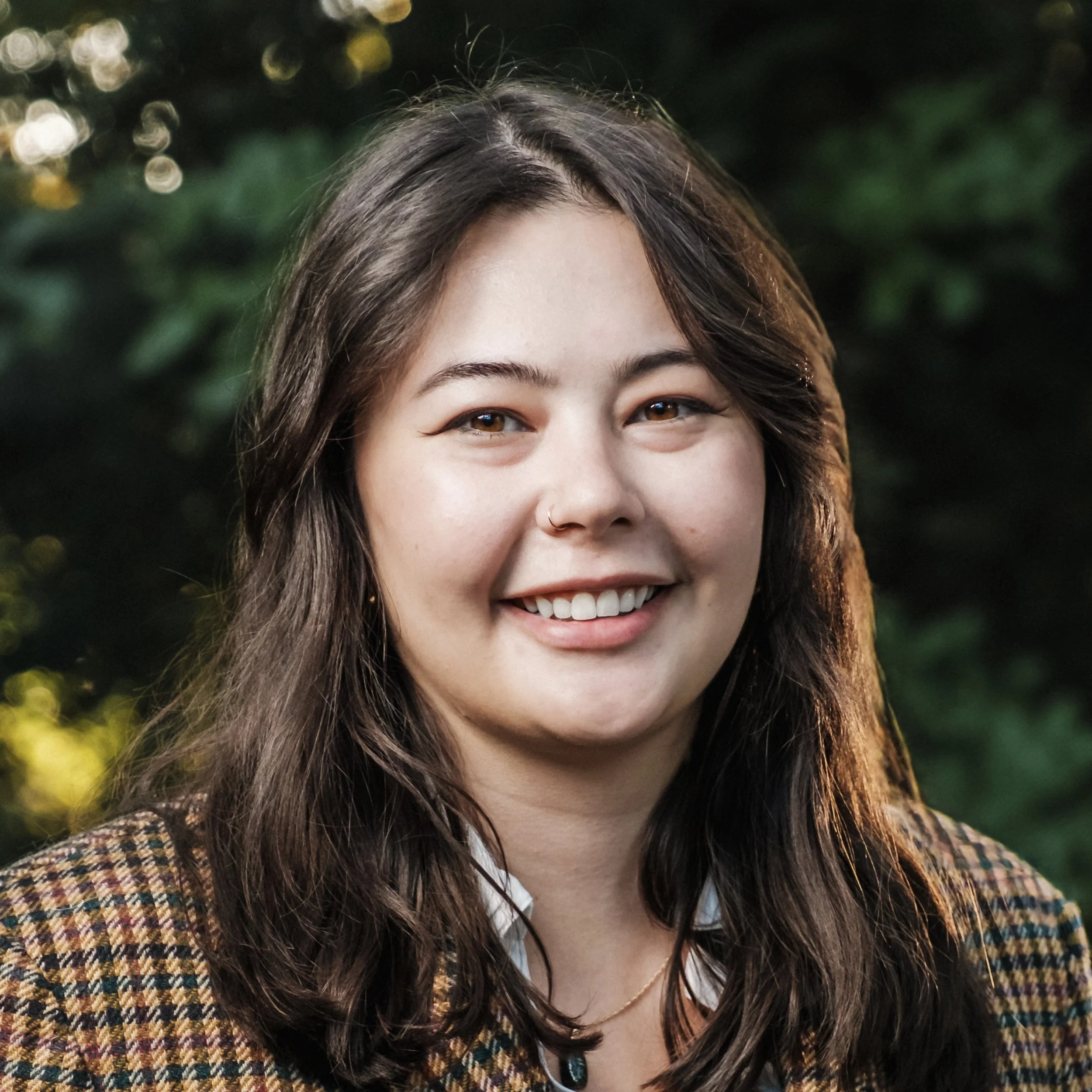 Close-up portrait of a smiling young woman with long dark hair, wearing a plaid jacket, outdoors with blurred green foliage in the background.