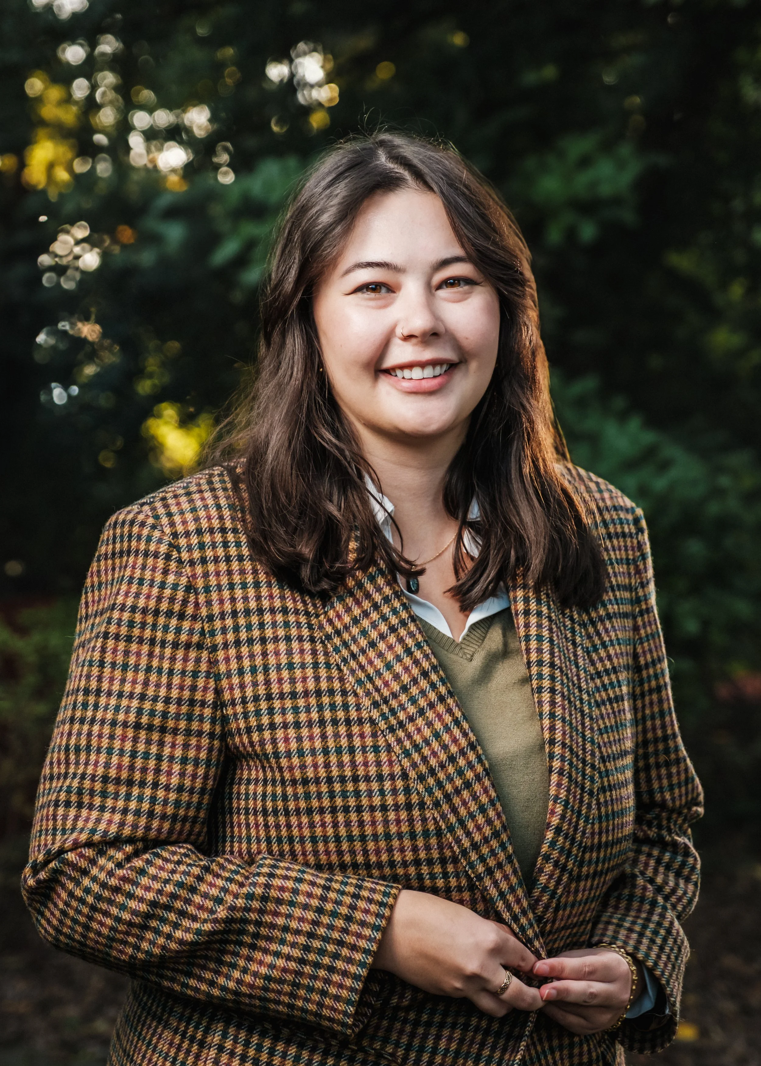 A young woman smiling outdoors, wearing a patterned blazer over a green sweater and white shirt, with greenery and sunlight in the background.