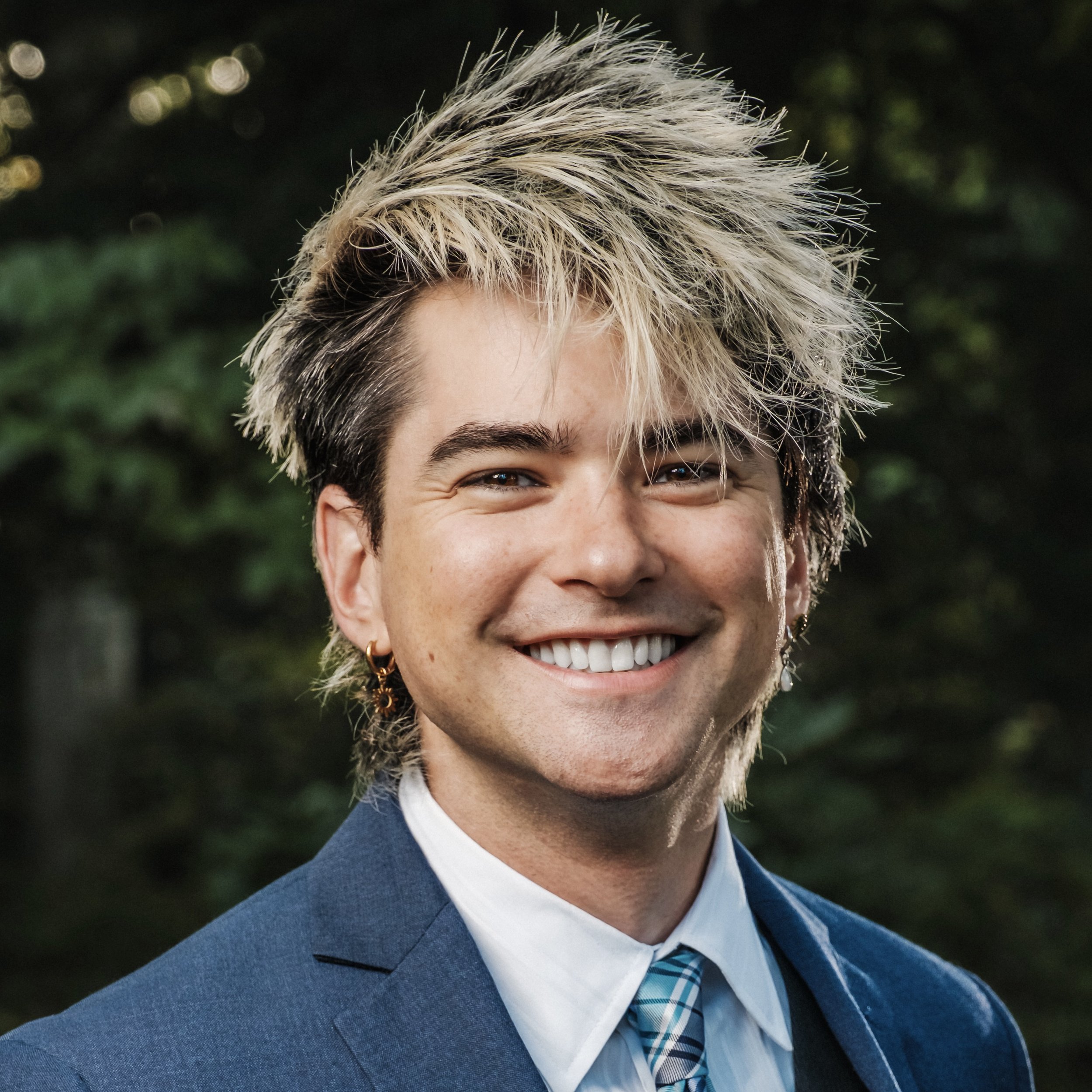 A young person with a big smile, short darker hair with blonde highlights, wearing earrings, a white shirt, a patterned tie, and a blue blazer, standing outdoors with blurred green trees in the background.