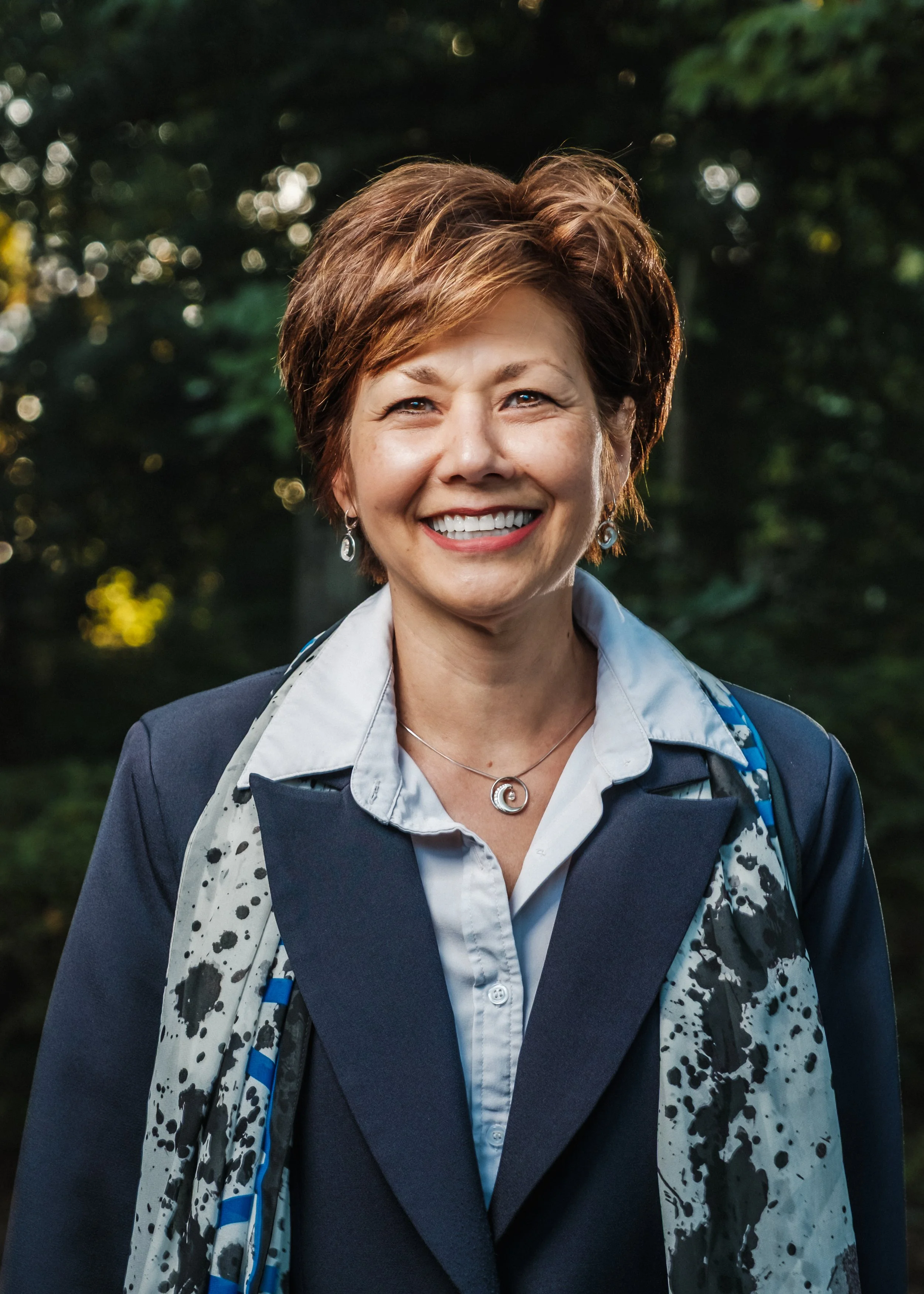 A woman with short, reddish-brown hair smiling outdoors in a wooded area, wearing a navy blazer, a white shirt, a patterned scarf, and jewelry.