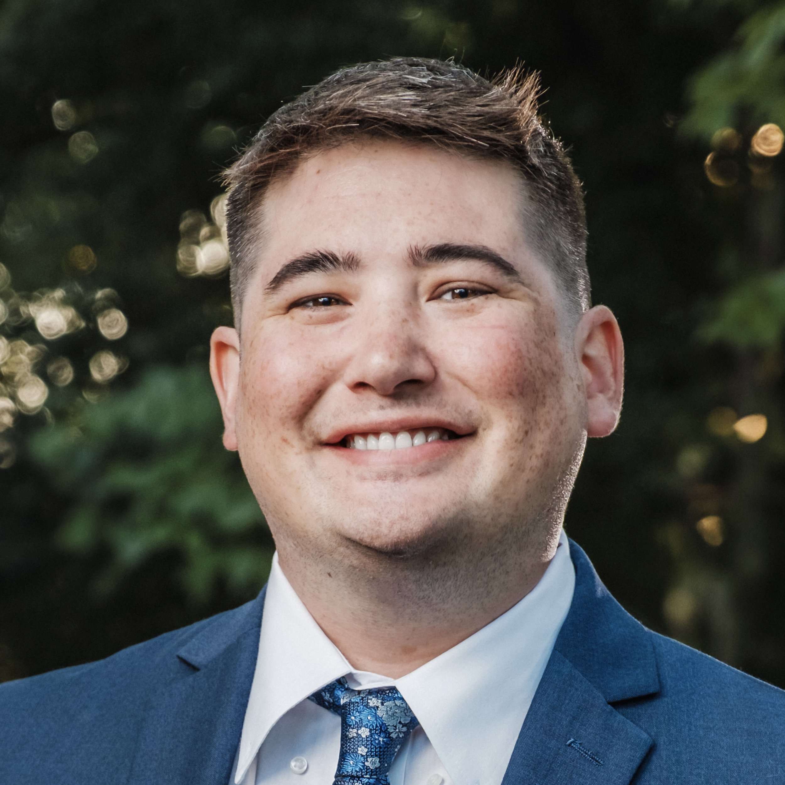 A smiling man in a blue suit, white shirt, and patterned blue tie outdoors with blurred greenery in the background.