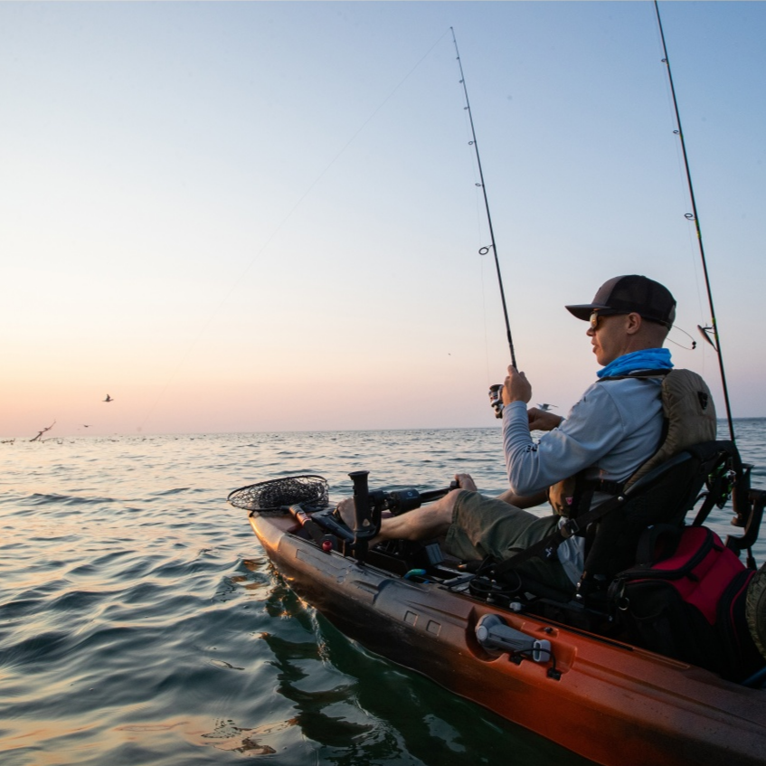Person fishing from a kayak on the water at sunset, with fishing rods and bird in the sky.