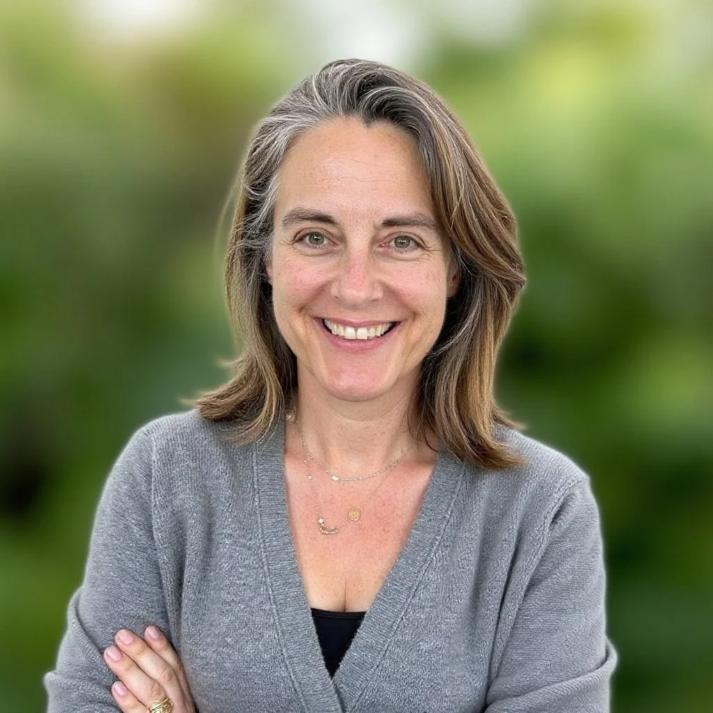 A woman with shoulder-length brown hair, smiling, wearing a gray cardigan, black top, gold necklace, and standing outdoors with a blurred green background.