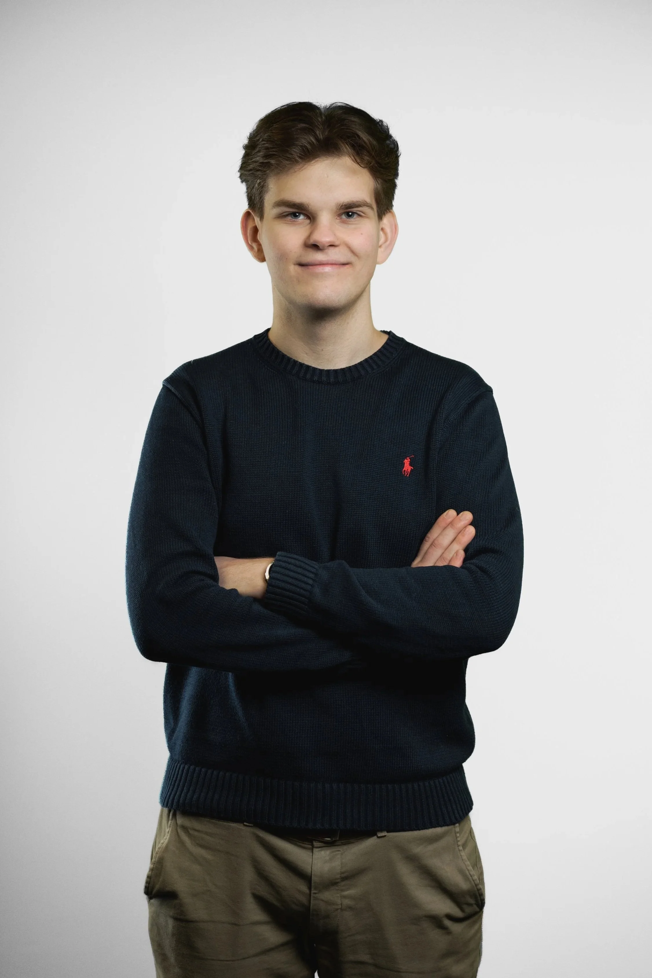 A young man with glasses and brown hair standing with arms crossed, wearing a dark sweater and khaki pants against a plain background.