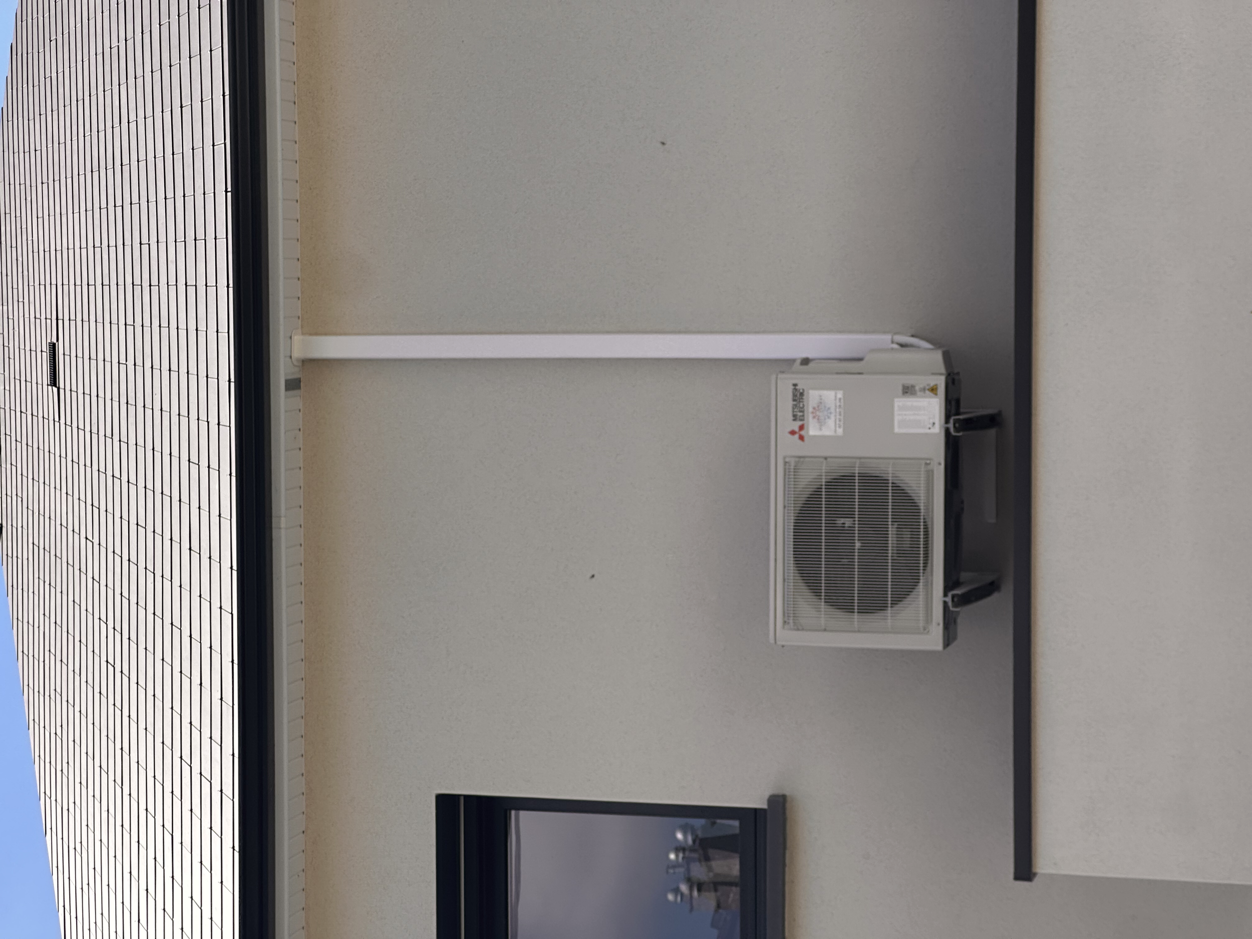 Un mur d'une maison avec une fenêtre noire, une unité de climatisation Mitsubishi Electric et une gouttière sur le toit.