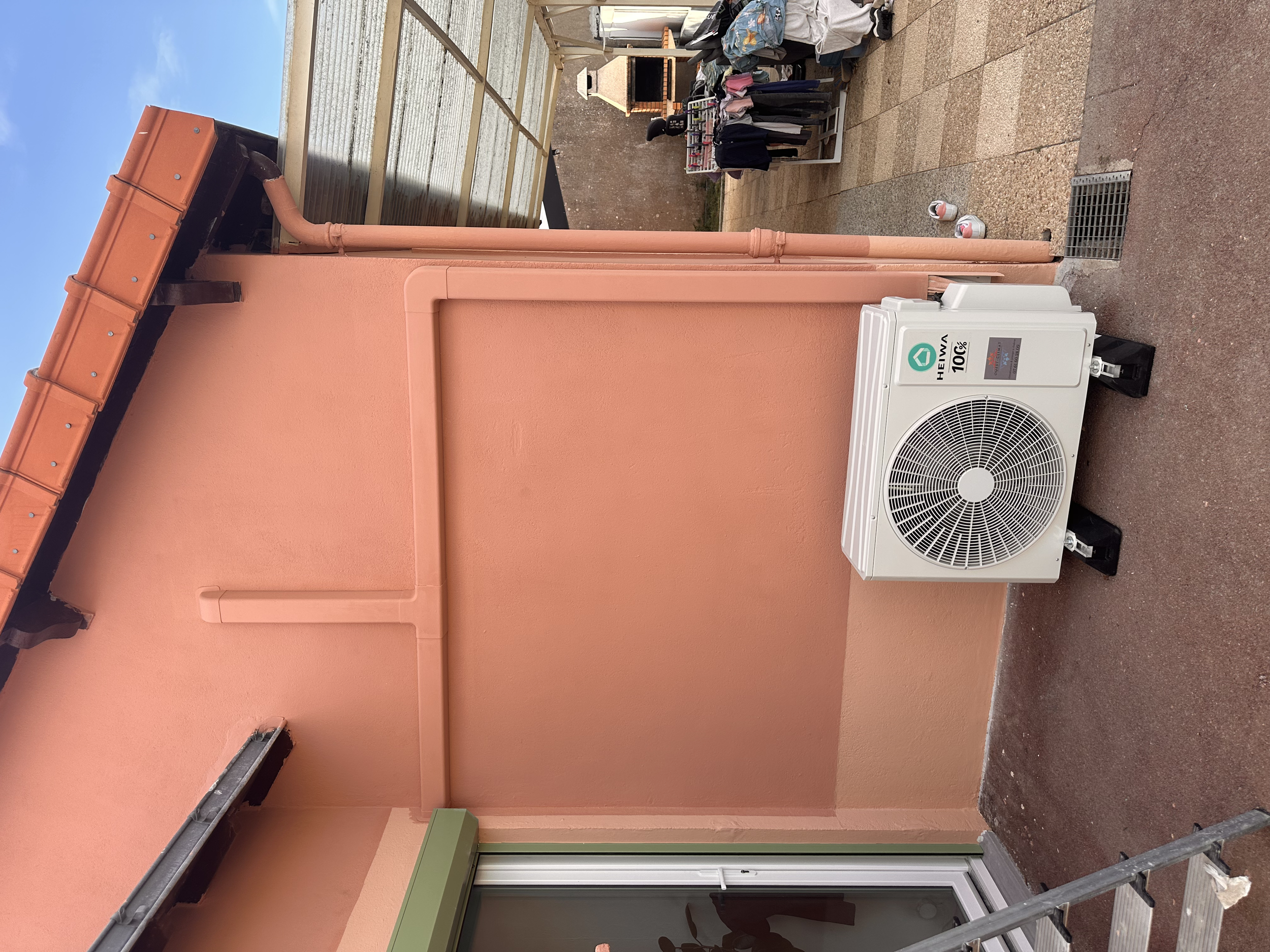 Climatisation extérieur HEIWA installé sur un mur rose avec un condenseur visible, dans une zone extérieure attenante à une maison, avec des vêtements suspendus pour sécher, un barbecue en pierre et des chaussures
