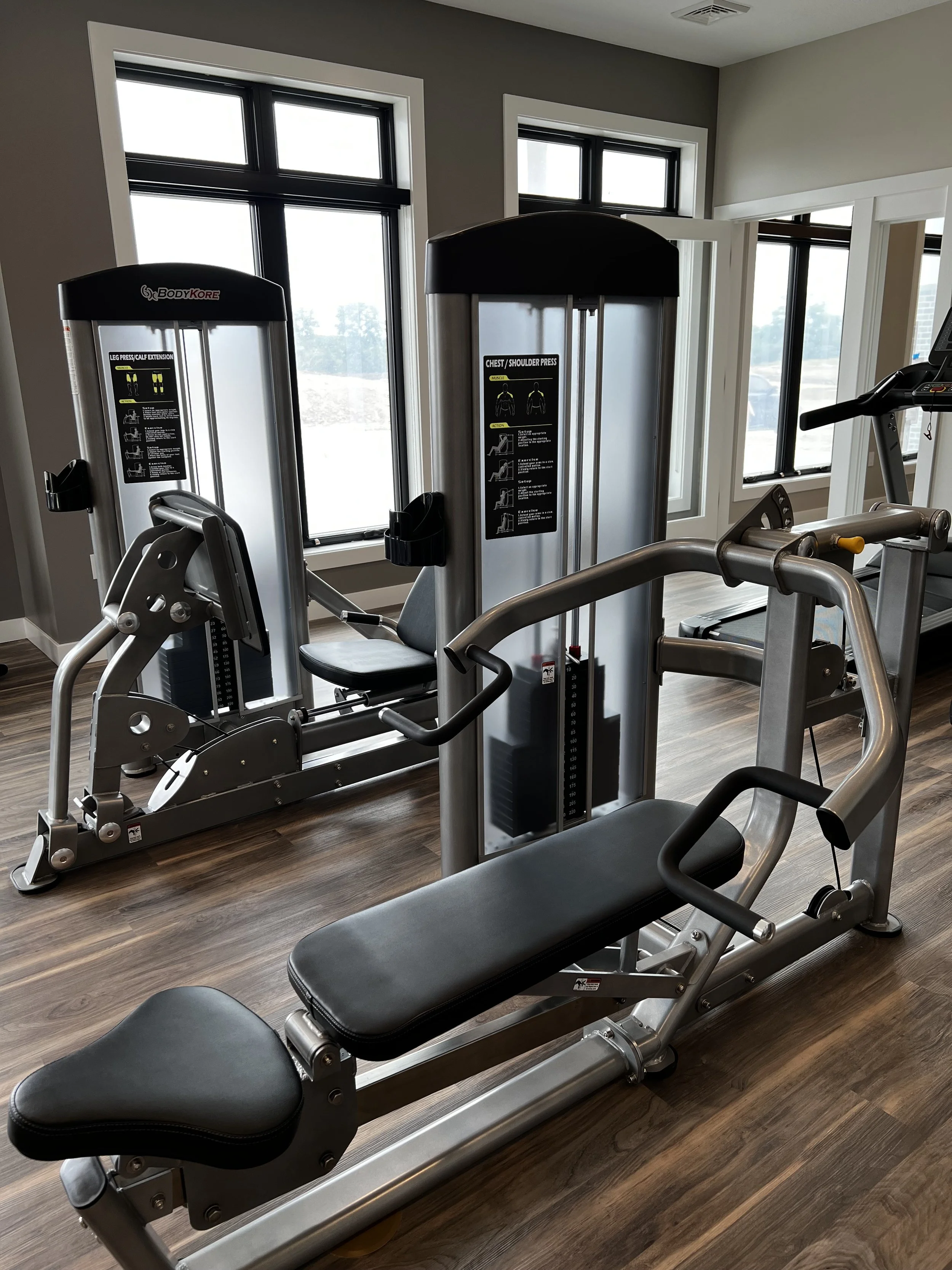 Fitness gym equipment including a leg press and chest/shoulder press machine, with hardwood floors and windows letting in natural light.