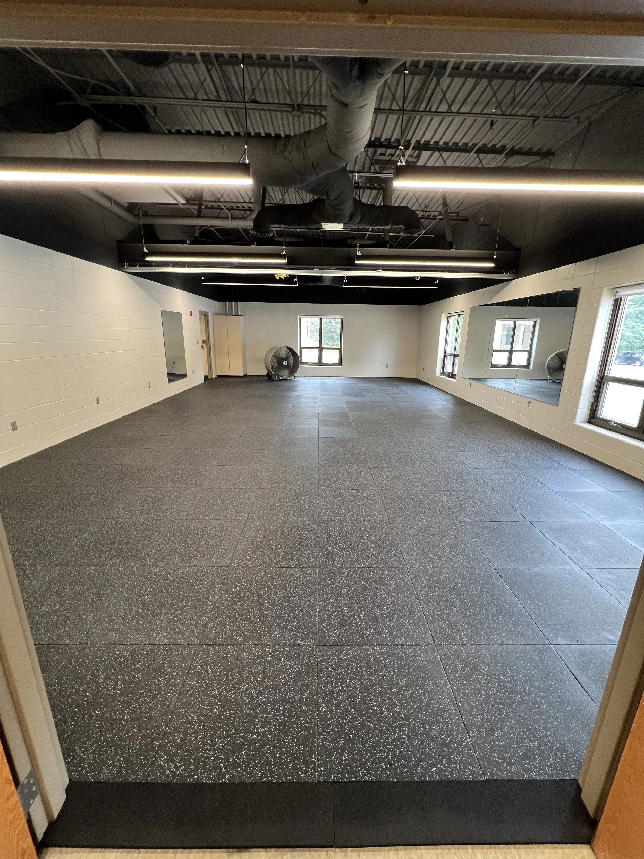 Empty fitness or dance studio with large mirrors on the wall, black ceiling with exposed ductwork, and a gray rubber floor. There are windows on two sides and a fan in the corner.