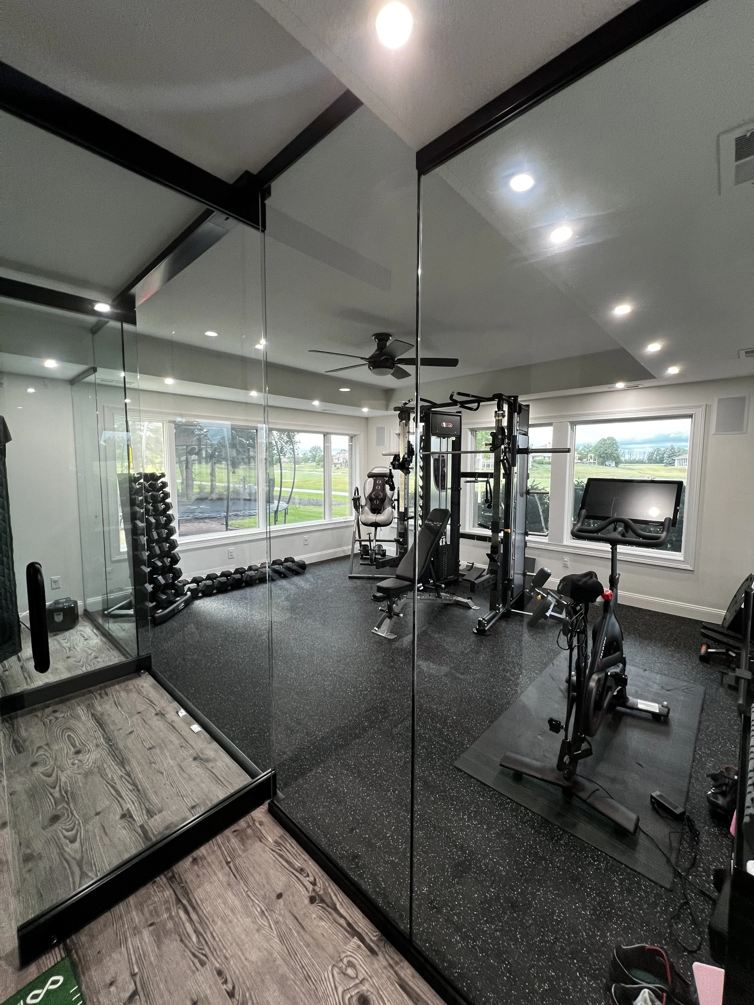 A home gym with black rubber flooring, exercise equipment including a stationary bike, a weight machine, a gym bench, and dumbbells. Large windows show a green outdoor landscape.