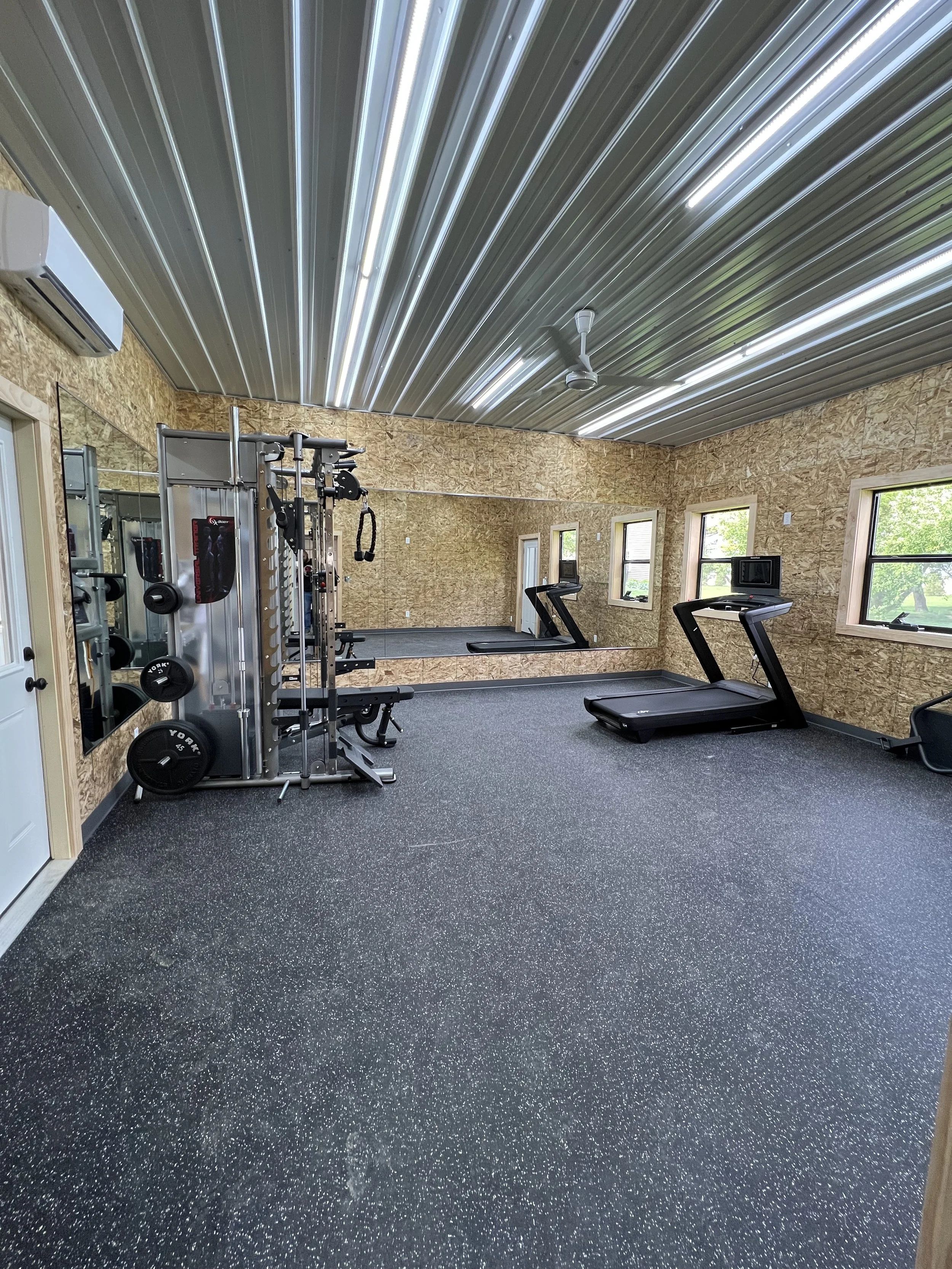 Home gym with workout equipment including a weight machine, treadmill, and mirror, with four windows in the walls and a metal ceiling.
