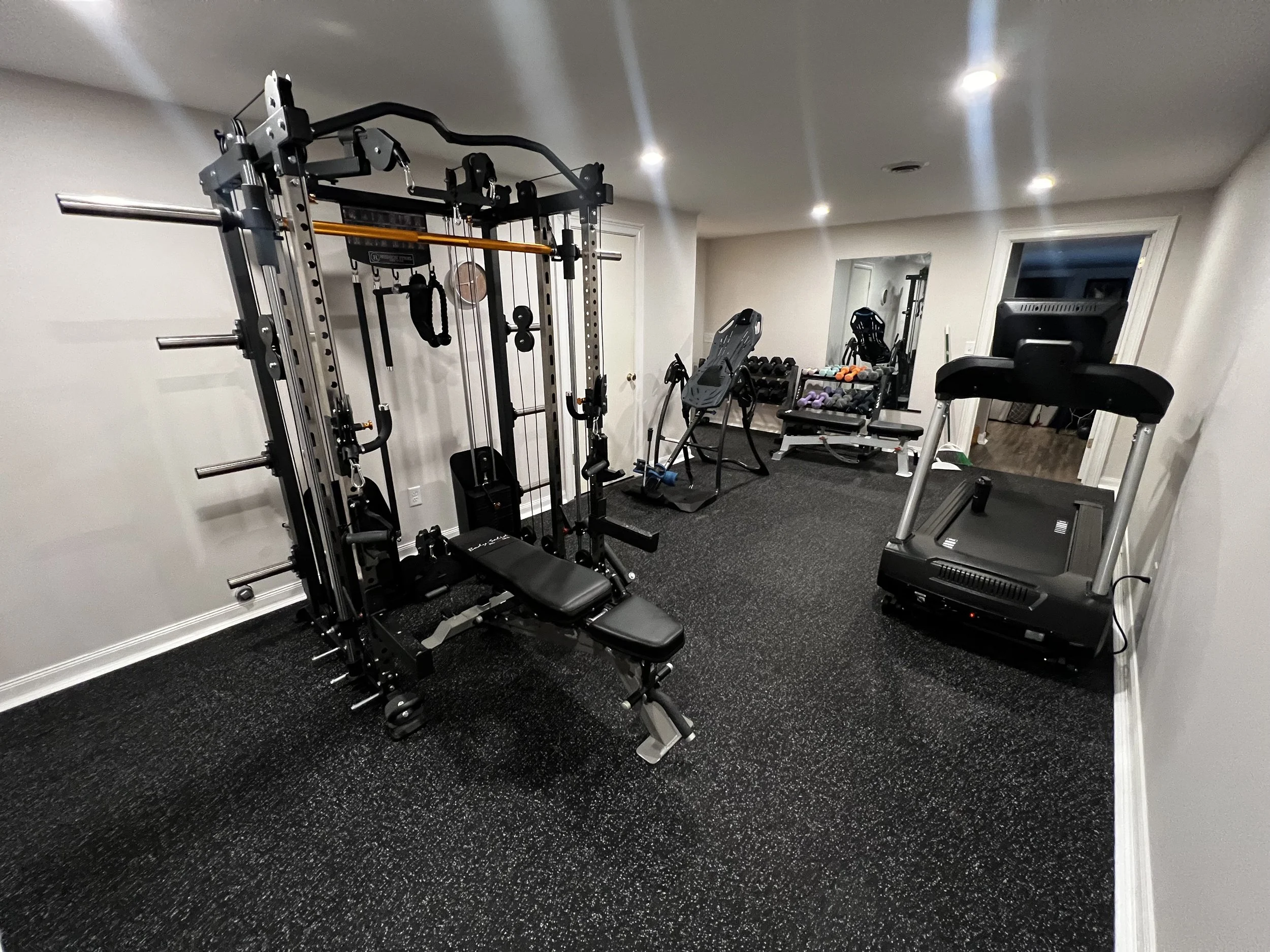 Home gymroom with various fitness equipment including a multi-function weight machine, treadmill, exercise bike, and free weights on racks, with a mirror on the back wall.