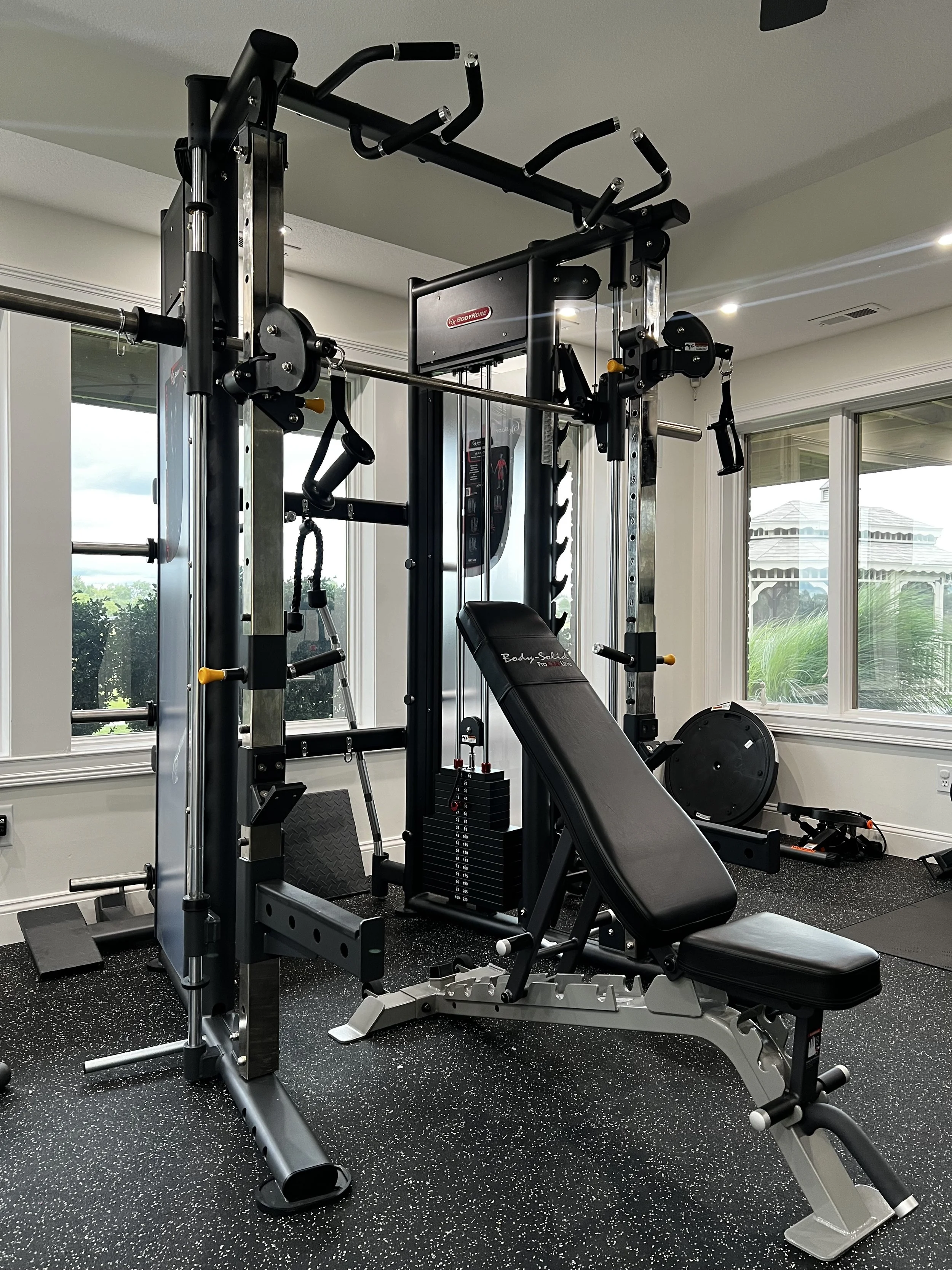 Home gym with a multi-station workout machine, adjustable bench, weight plates, and additional exercise equipment near large windows.