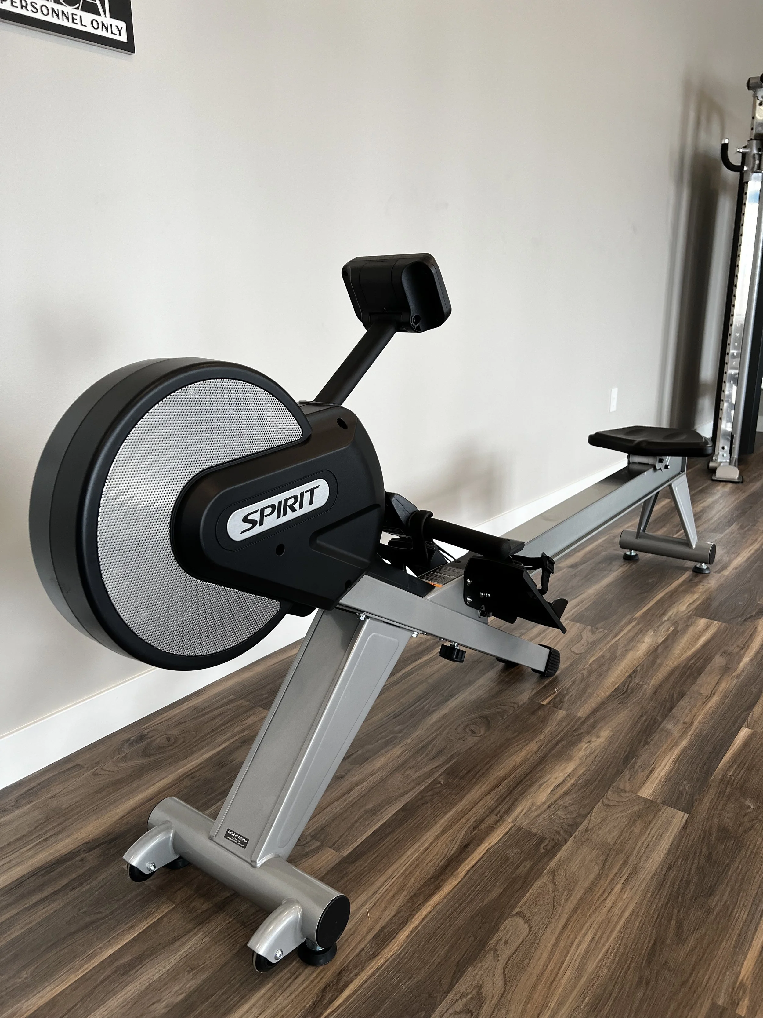 A rowing machine in an indoor gym with wooden flooring and a plain white wall.