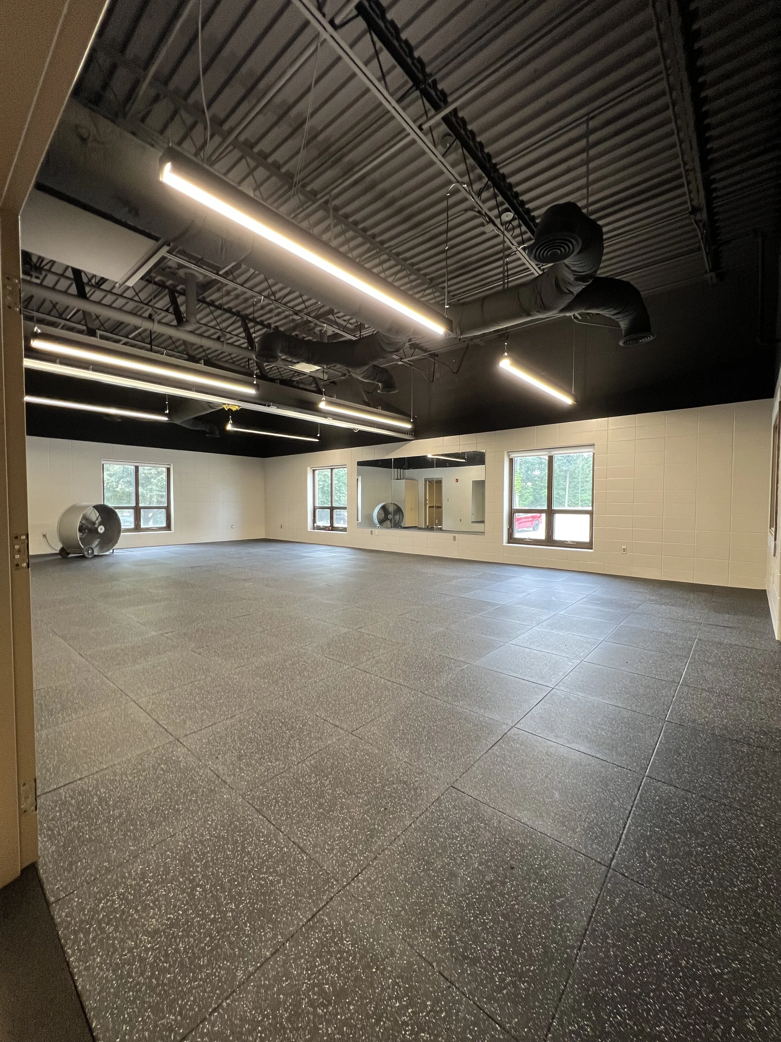 Empty fitness or dance studio with black flooring, large mirrors on one wall, three windows, and black ceiling with exposed ducts and lighting.