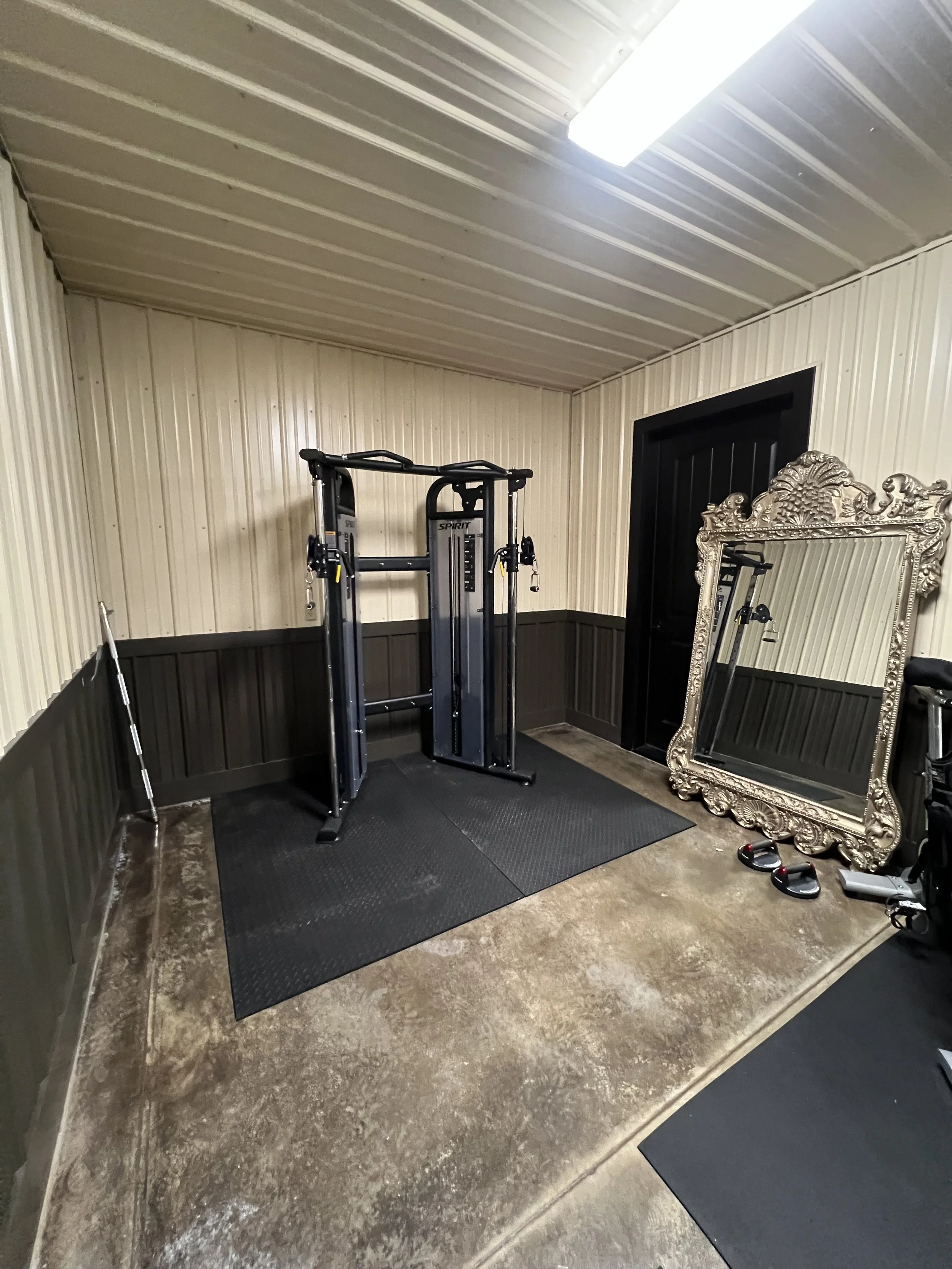 Home gym corner with a black exercise cable machine, a large ornate mirror, a pair of weightlifting gloves, a black doormat, and a black door, with beige and brown paneled walls and a fluorescent ceiling light.