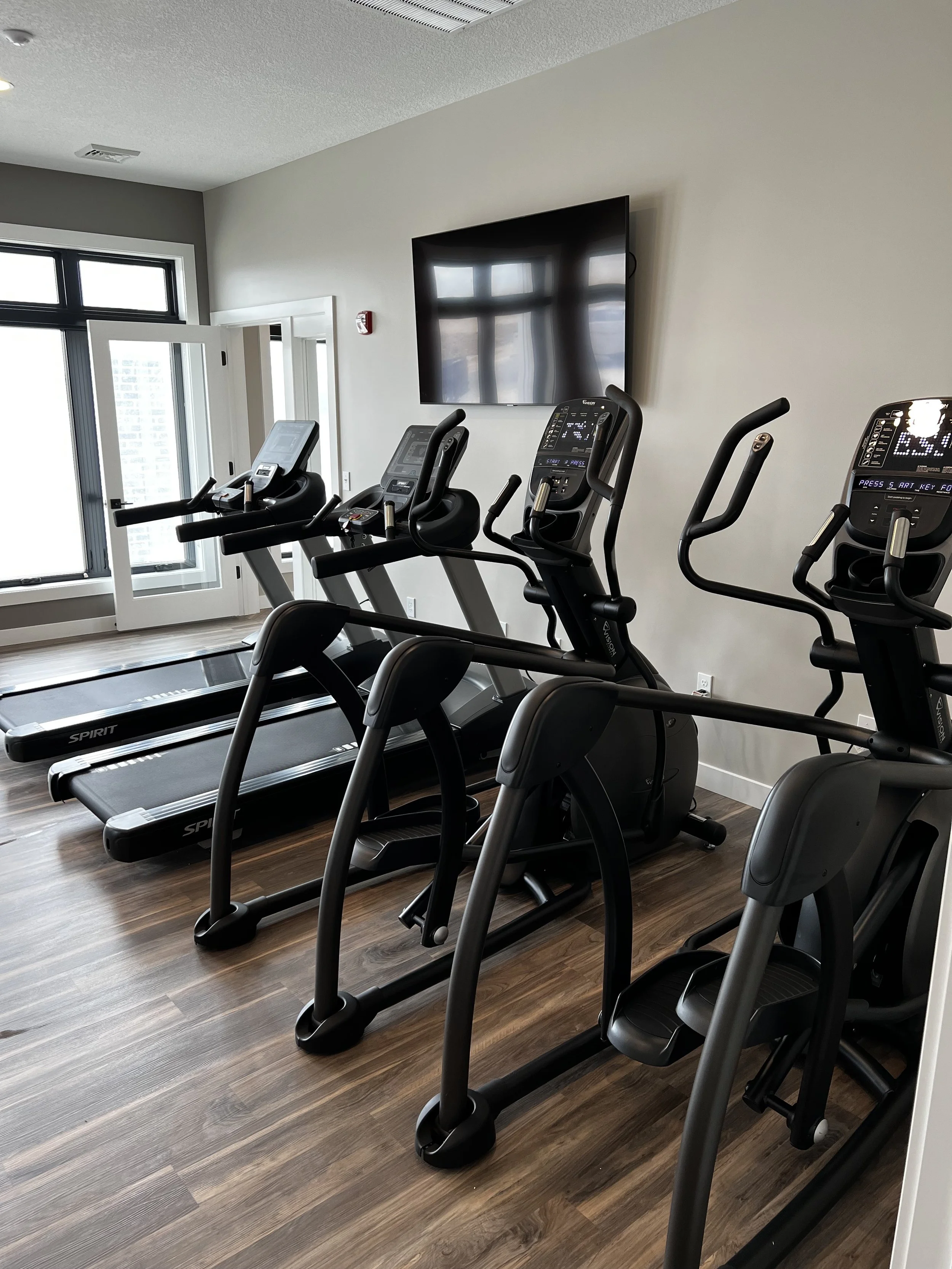 A small gym room with four black exercise machines, including treadmills and elliptical trainers, on a wooden floor, with a wall-mounted television and large windows letting in natural light.
