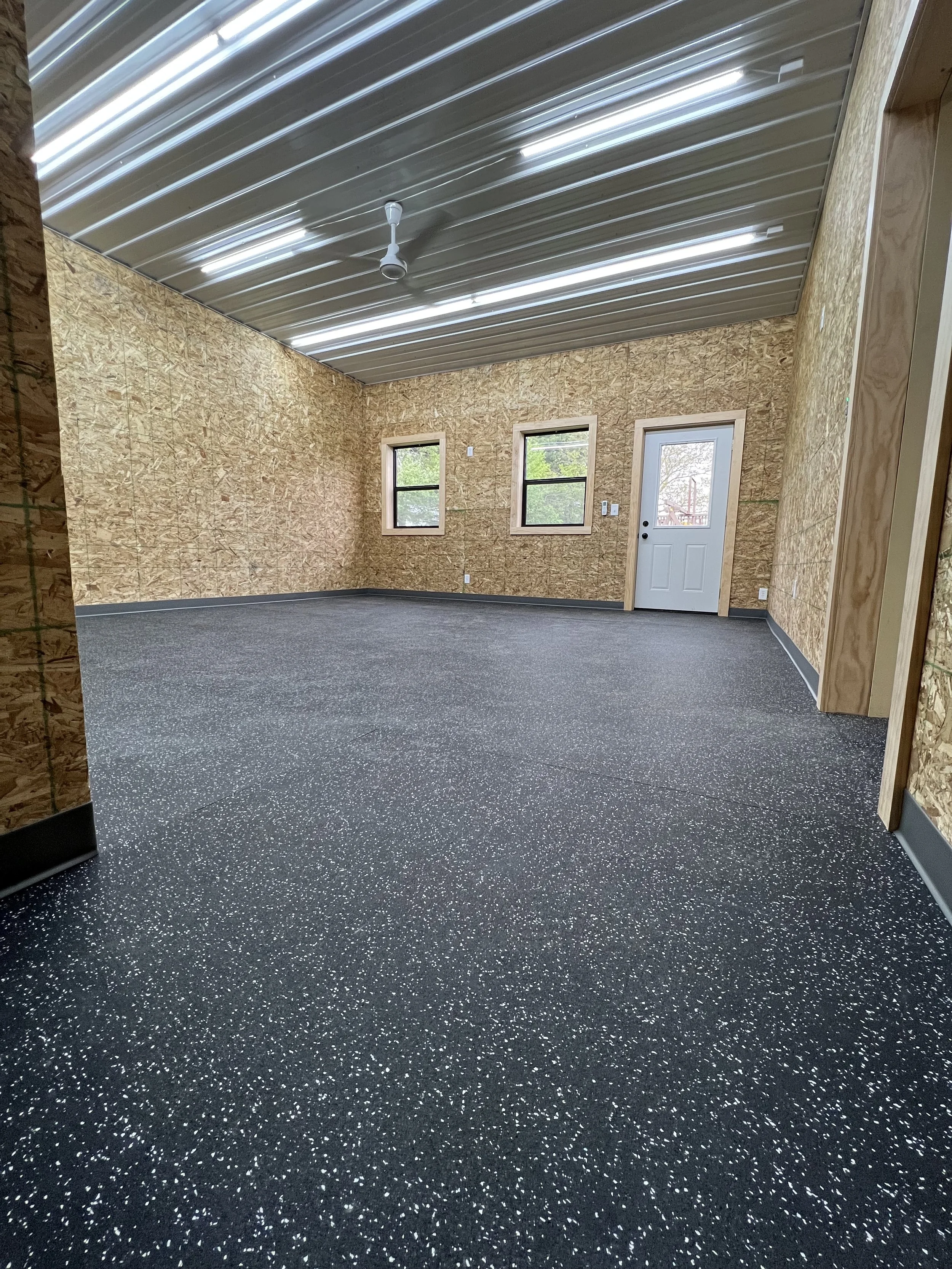 Unfinished indoor space with plywood walls, black speckled flooring, and a metal ceiling, featuring three windows and a white door.