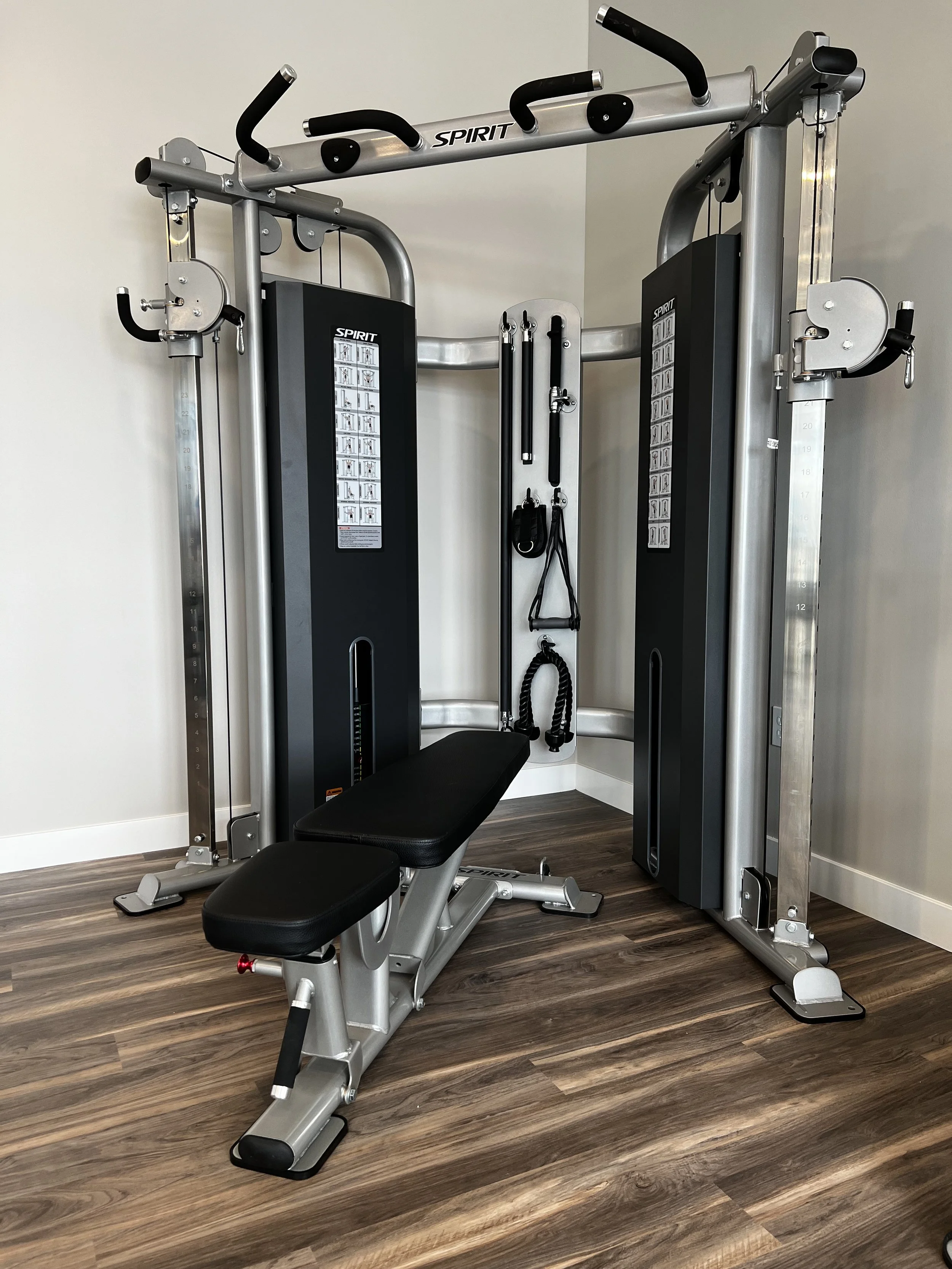 A cable machine gym equipment with adjustable pulleys, a padded bench, and resistance straps, labeled 'Spirit', set up against a plain wall with wood flooring.