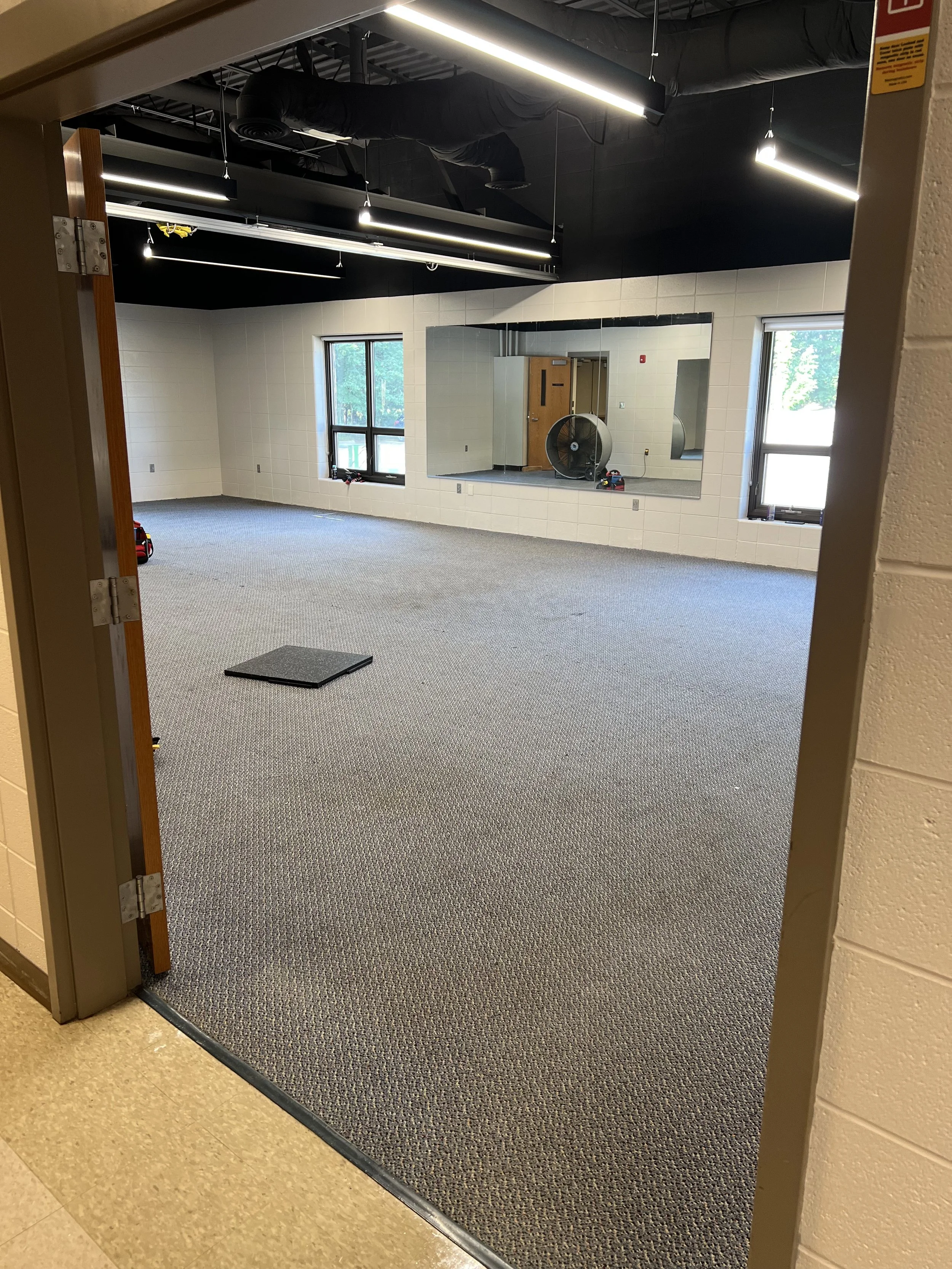 Empty room with a carpeted floor, two windows, a large mirror on the wall, and black ceiling with lighting fixtures and exposed ducts.