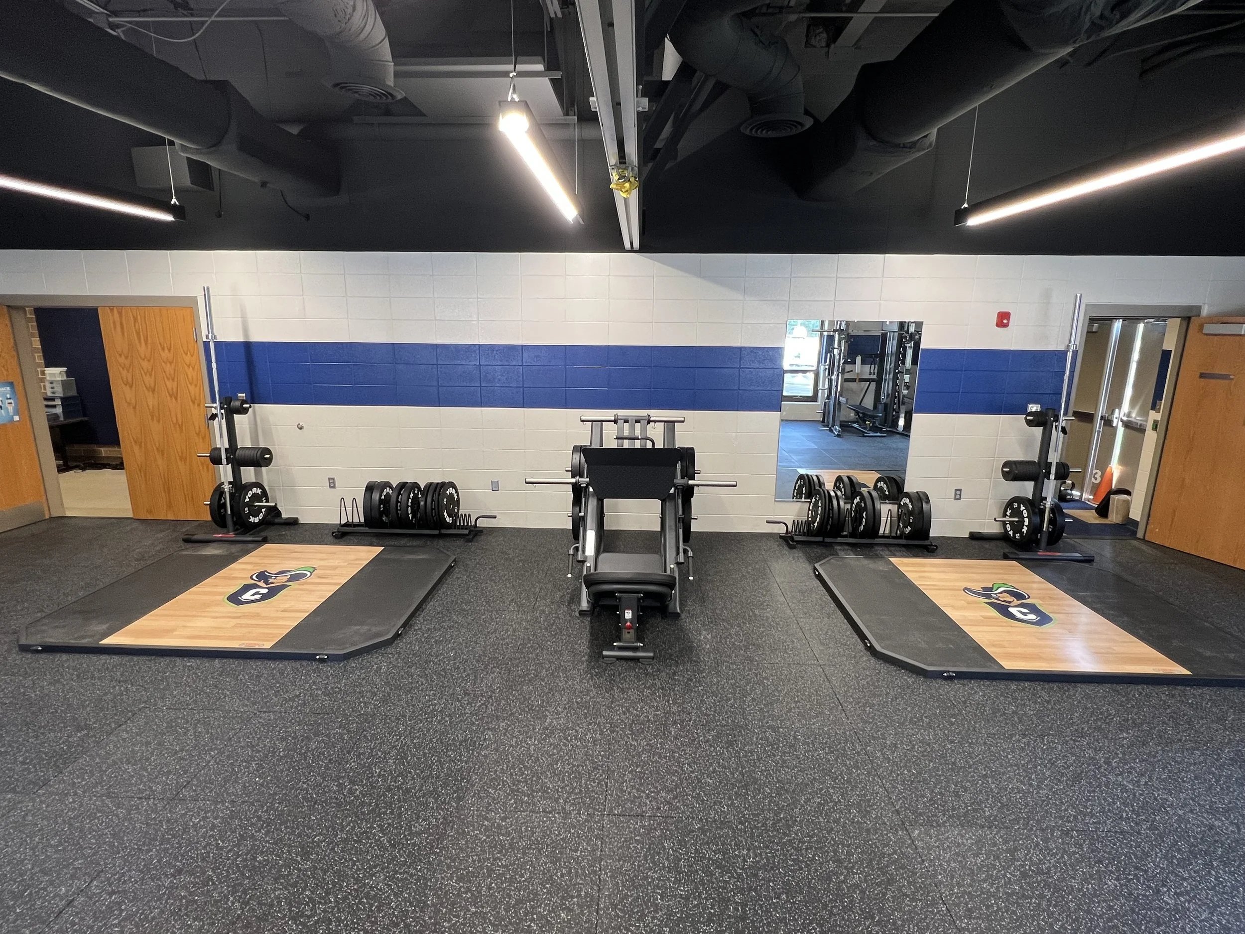 Empty strength training room with two weightlifting platforms, barbells, and weight plates, with a mirror on the wall and a door on each side.