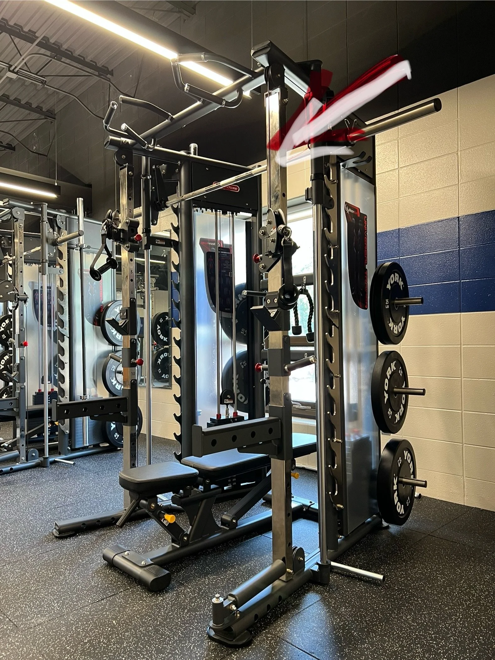 Weightlifting and strength training equipment in a gym, including a Squat rack, weight plates, and a bench, with mirrors and wall padding.