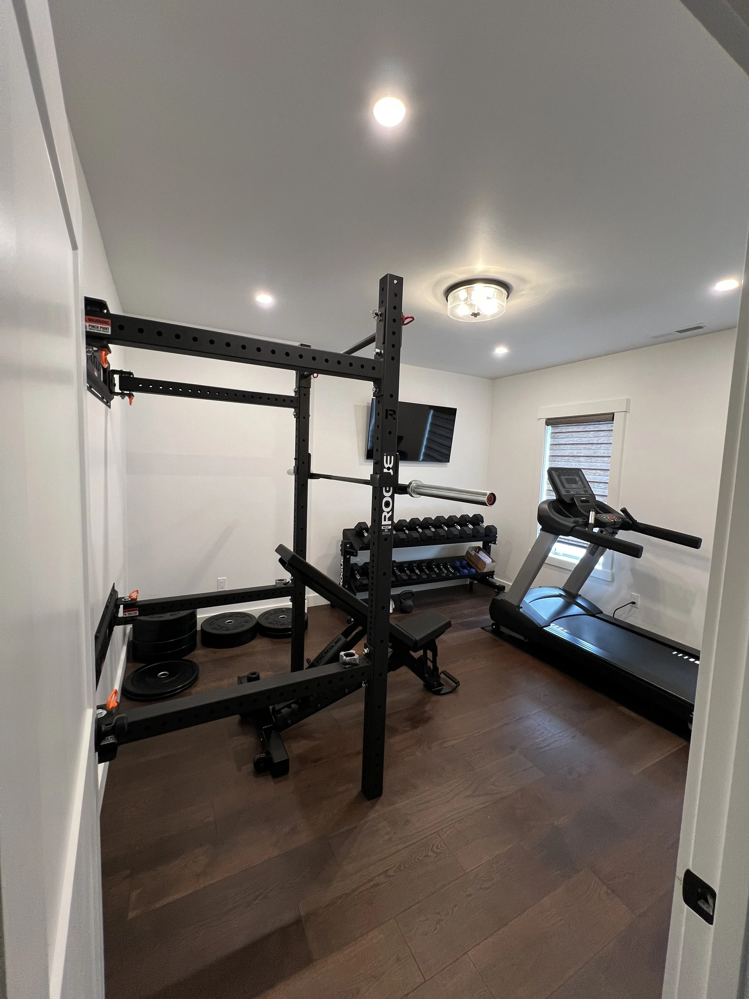 Home gym room with treadmill near window, black power rack with barbells, and a set of dumbbells on a rack, dark wood flooring, white walls, ceiling lights, and a mounted TV.