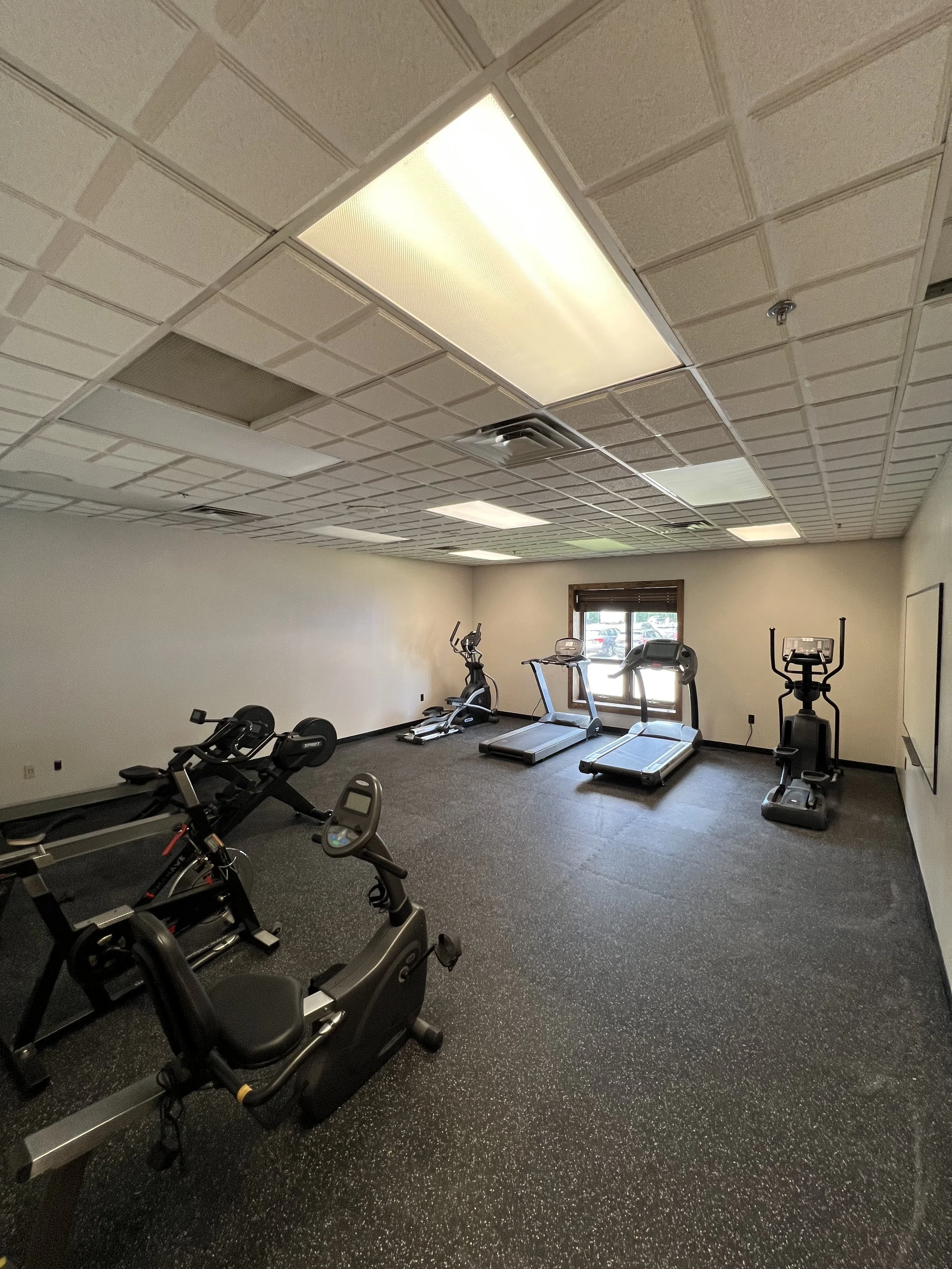 Empty small gym room with exercise equipment including a treadmill, stationary bikes, and elliptical machines, with a window on the back wall.