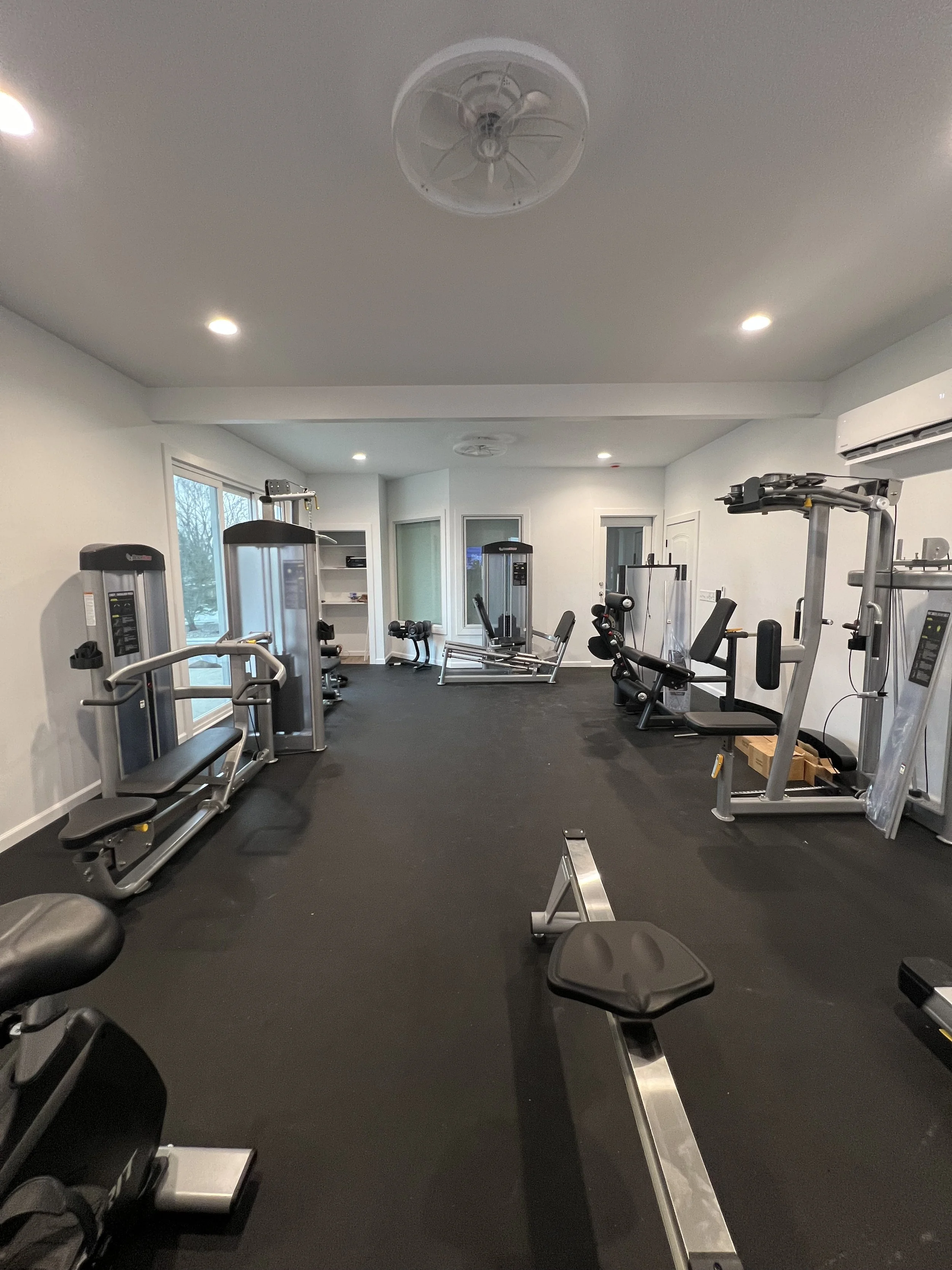 Empty gym room with various exercise equipment including weight machines, benches, and cardio machines, with large windows and white walls.