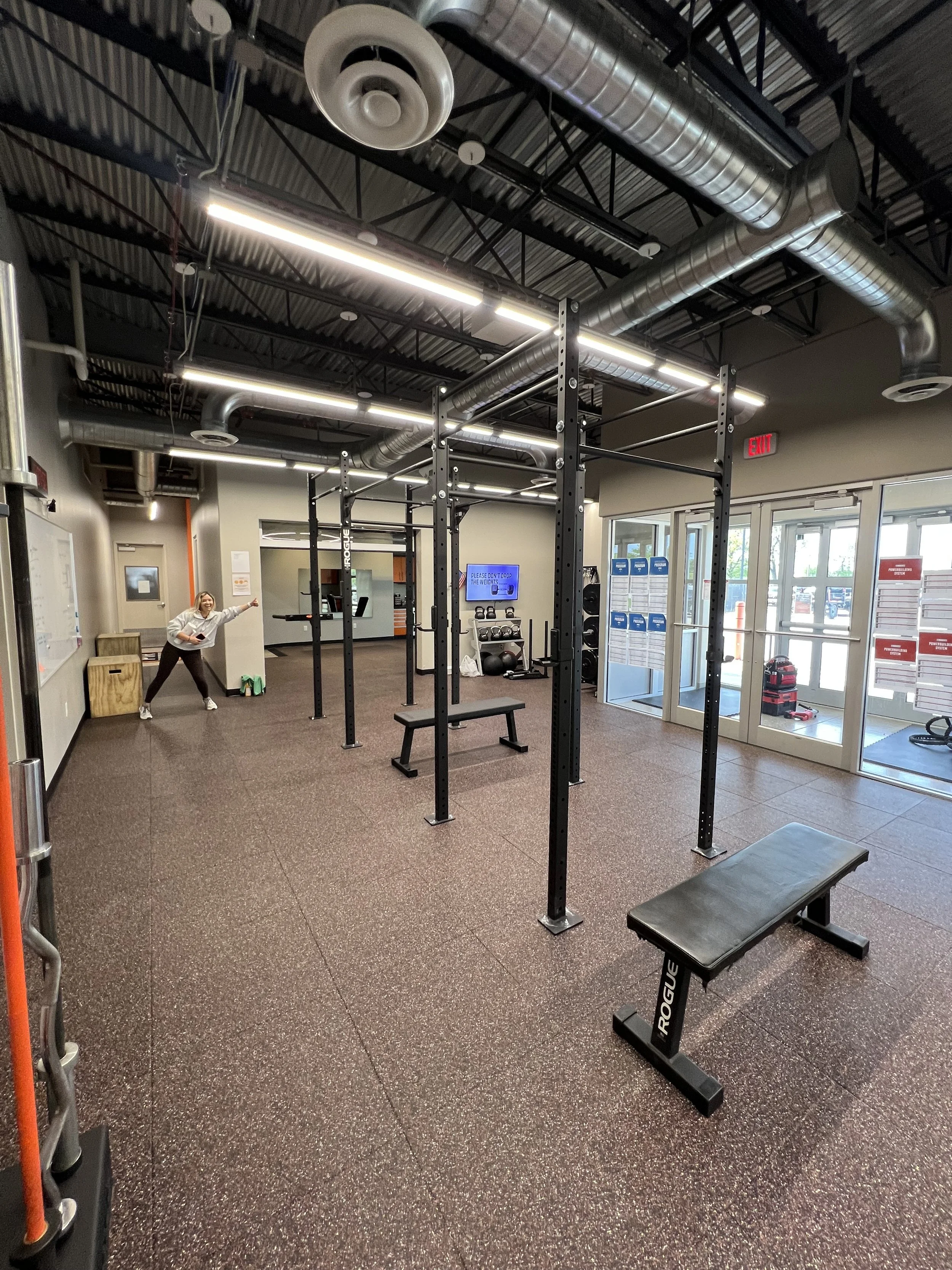 Empty gym with black metal pull-up and squat racks, benches, and workout equipment. A woman is standing in the background near a whiteboard, smiling. Large windows and bright ceiling lights illuminate the space.