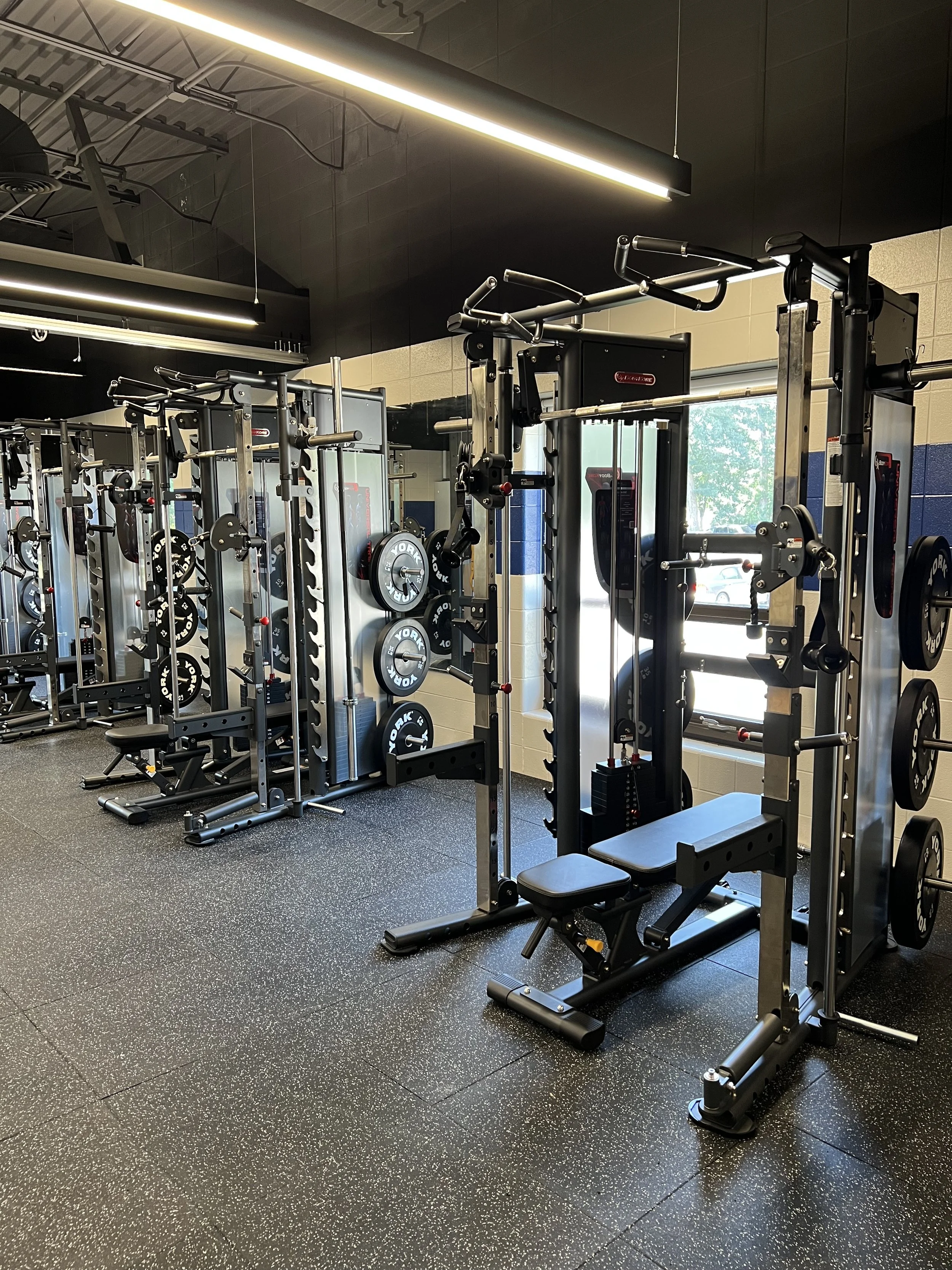 Interior view of a gym with various weightlifting equipment, including squat racks, barbell weights, and benches.