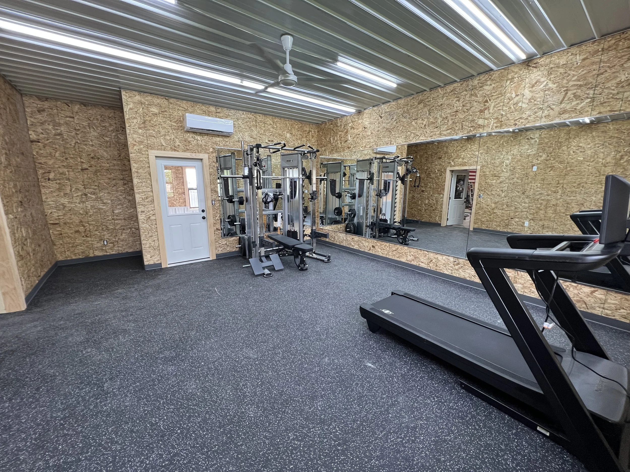 Empty gym room with wood-paneled walls and a large mirror, featuring exercise equipment such as a treadmill, weight machines, and a door leading outside.