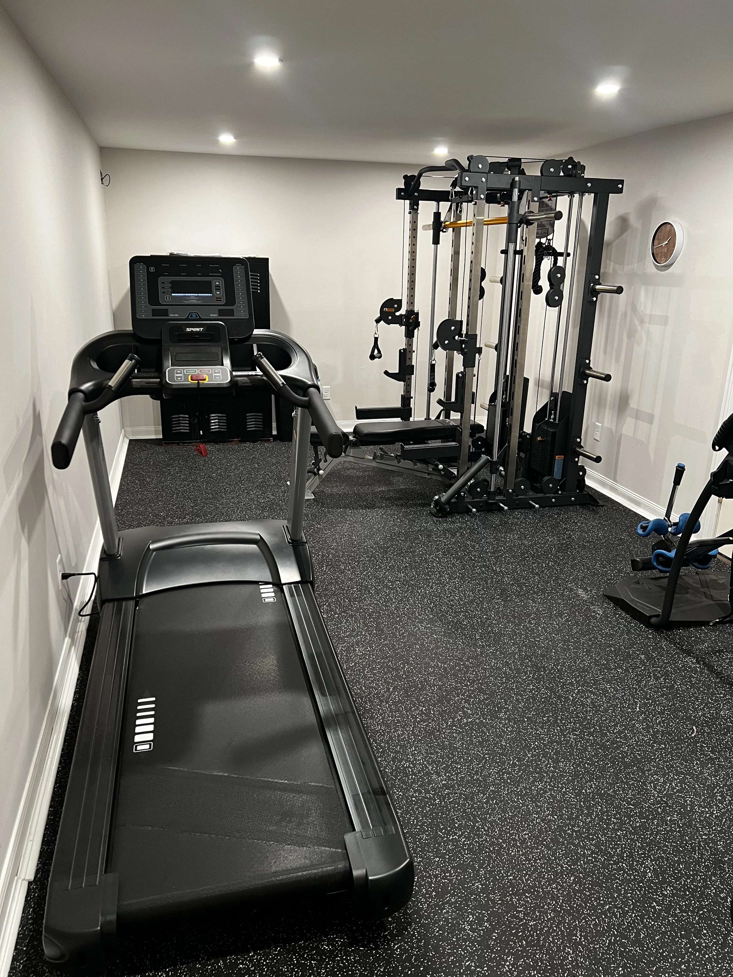 Home gym with treadmill, weight machine, small clock on wall, and exercise accessories.