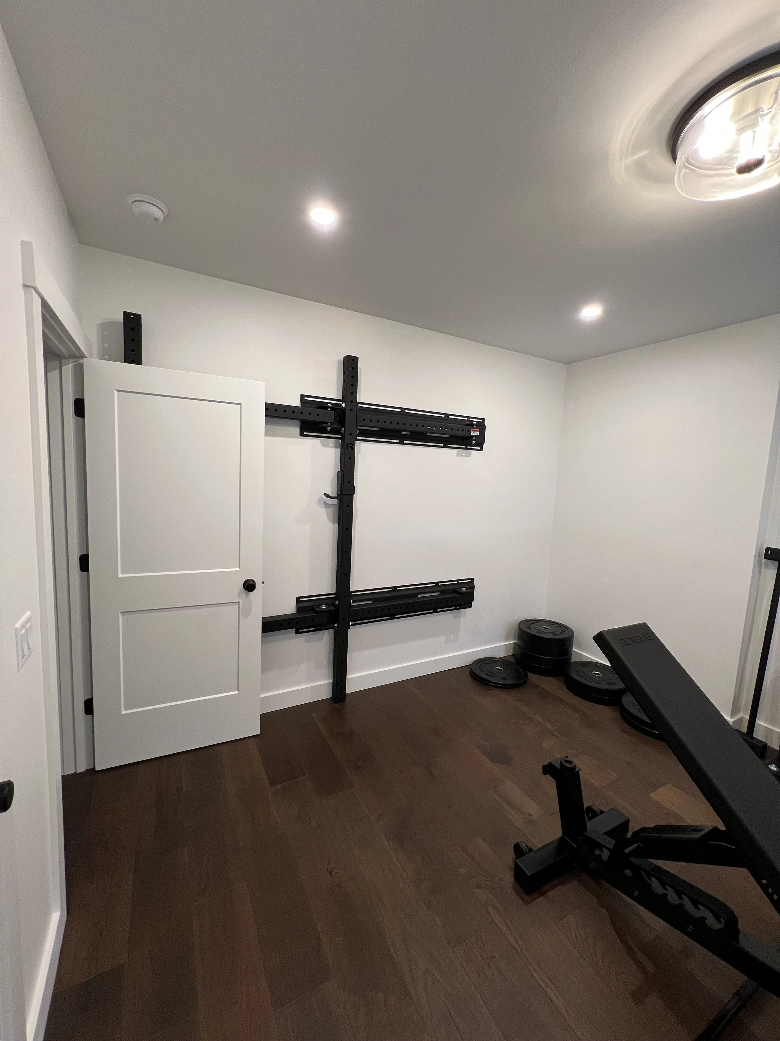 Empty home gym room with a wall-mounted pull-up station, a small door, and a pile of weight plates on the floor.