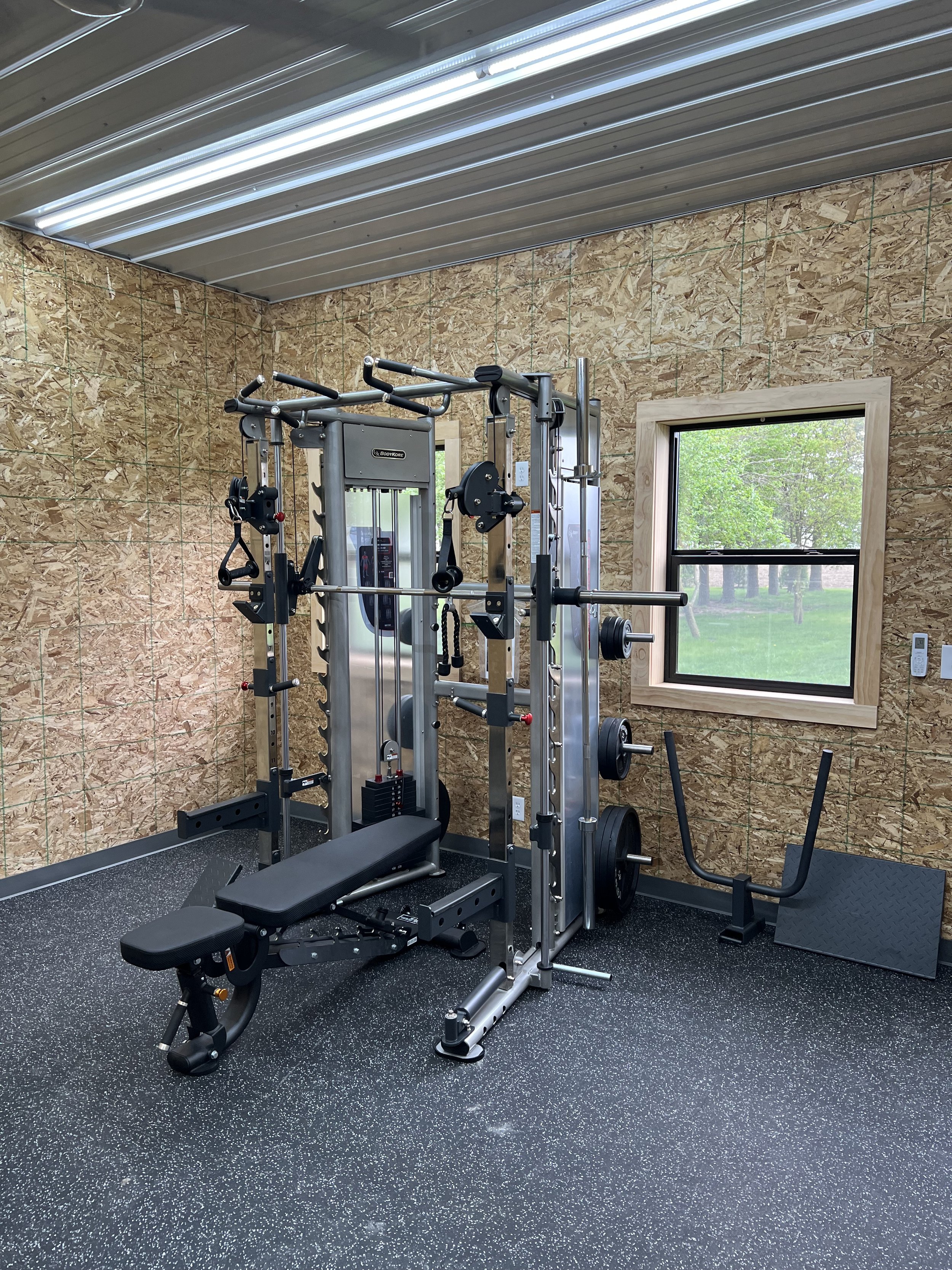 Home gym with cable weight machine, adjustable bench, and a small sit-up bench, set against wood panel walls and a window showing grass and trees outside.