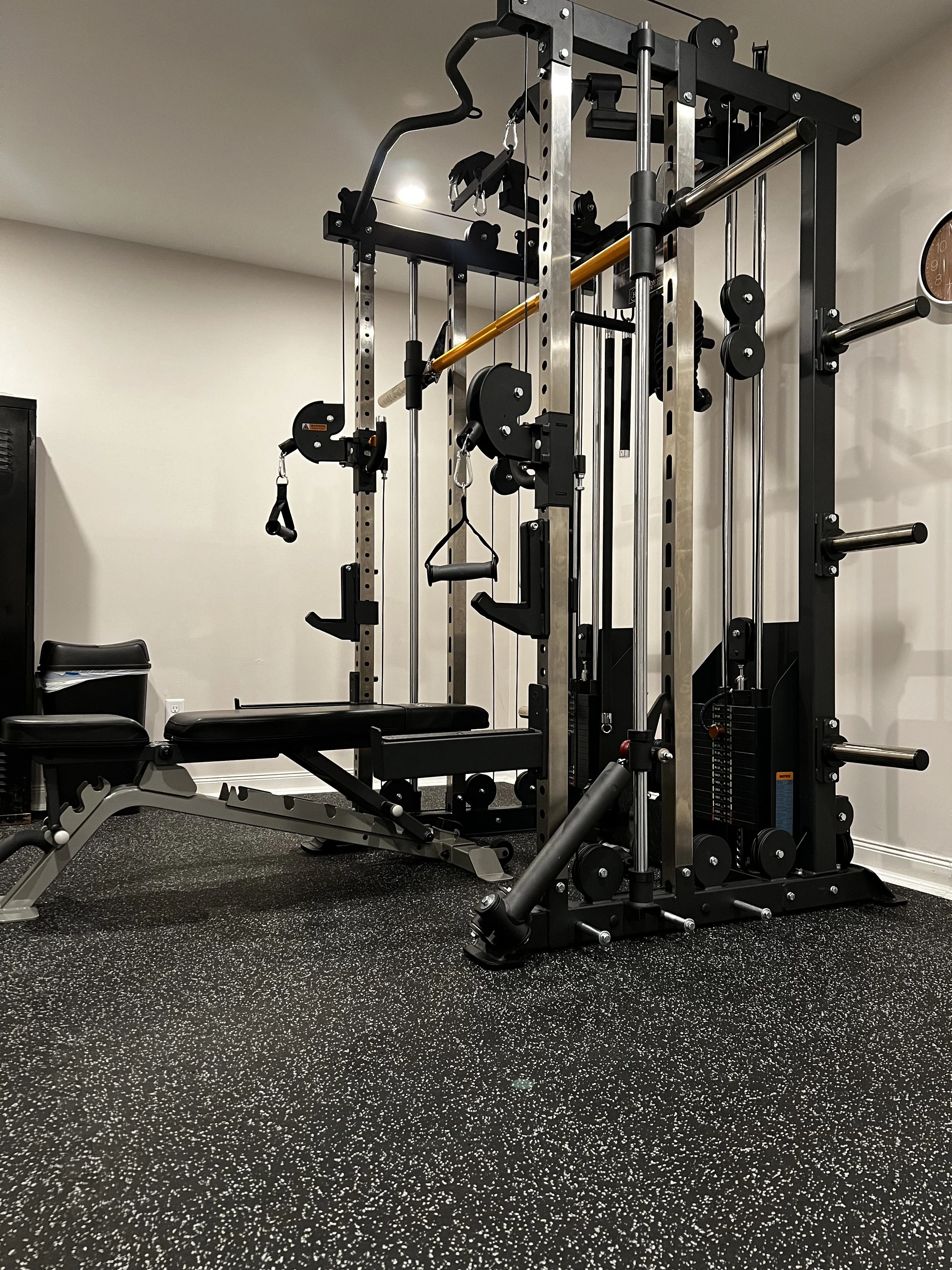 A home gym with a multi-function weight machine, black and metal frame, bench, and weight plates, situated on a speckled black flooring with a white wall background.