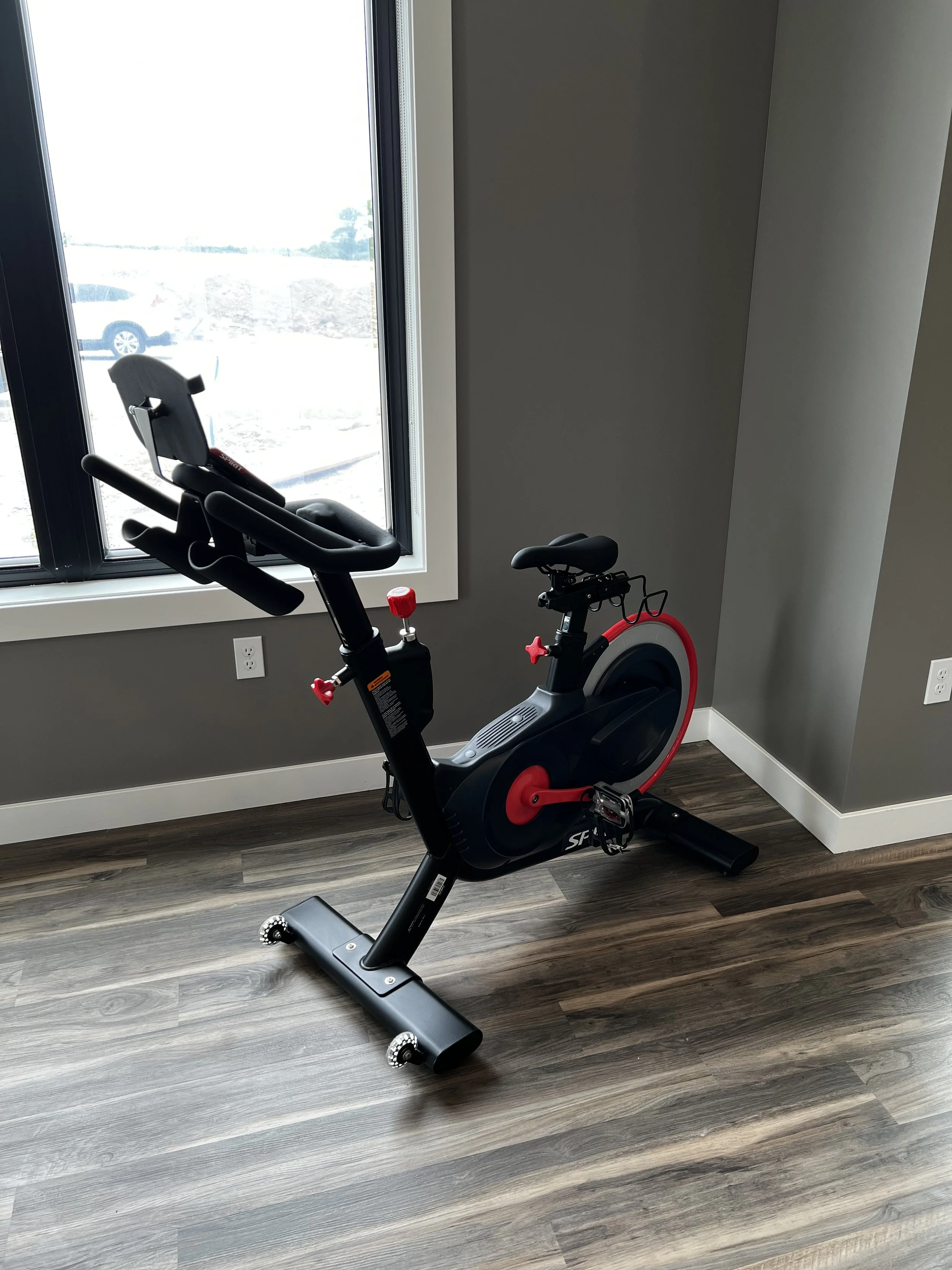 Indoor room with a stationary exercise bike near a window, wood laminate flooring, and gray walls.