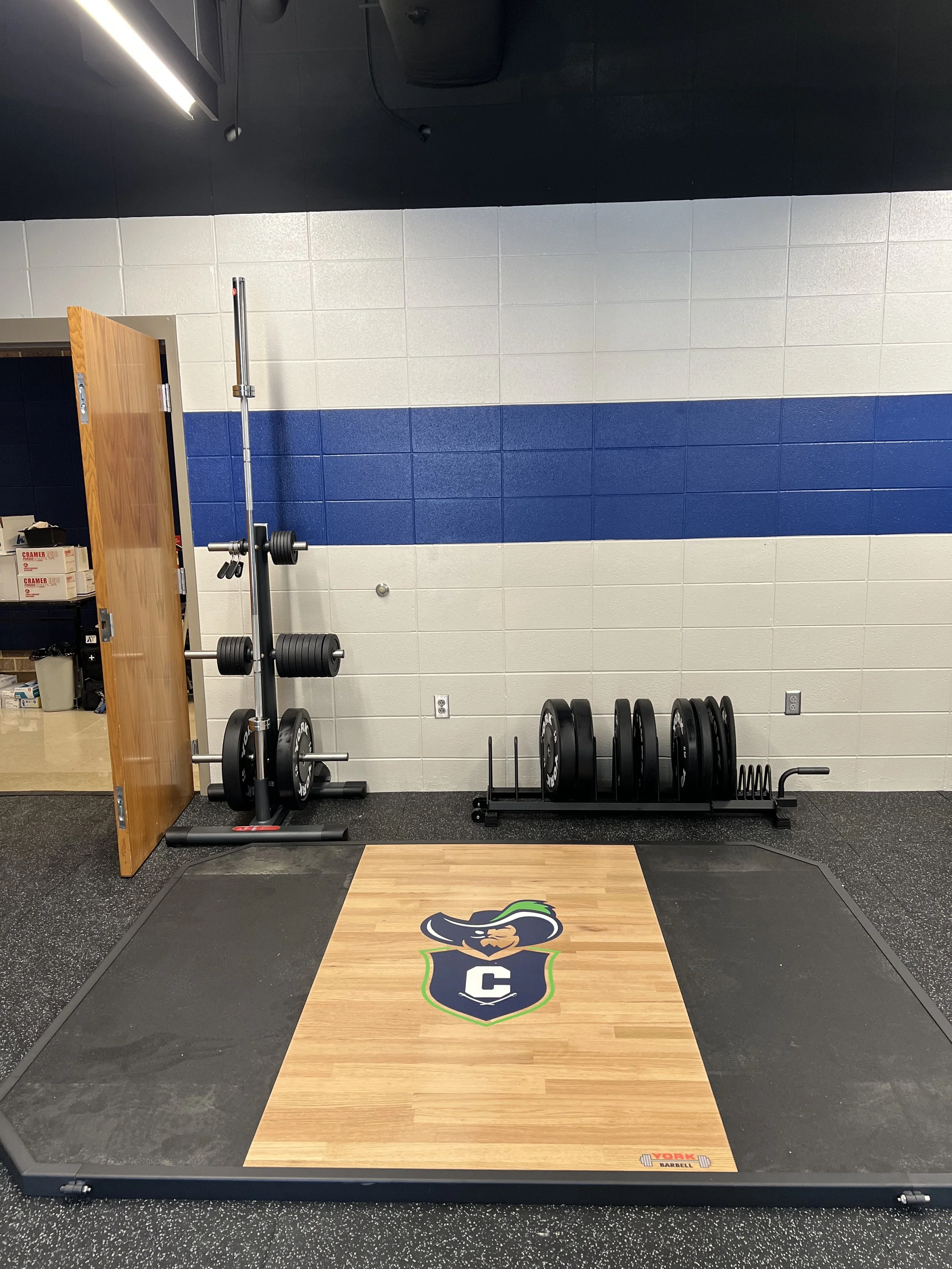 Empty weightlifting area with a large platform, weight plates on a rack, and a barbell with weights. The platform features a logo of a hawk or eagle with a shield and the letter 'C'.