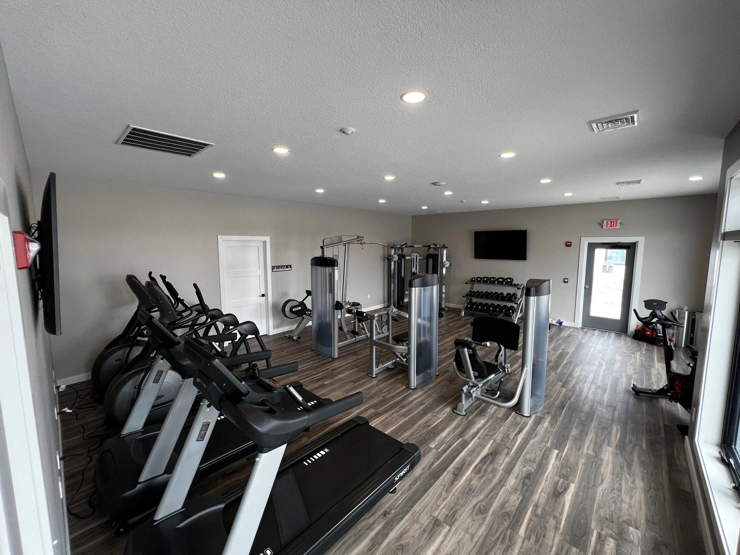 Empty gym with cardio machines, strength training equipment, a television, and large windows, with hardwood floors and gray walls.
