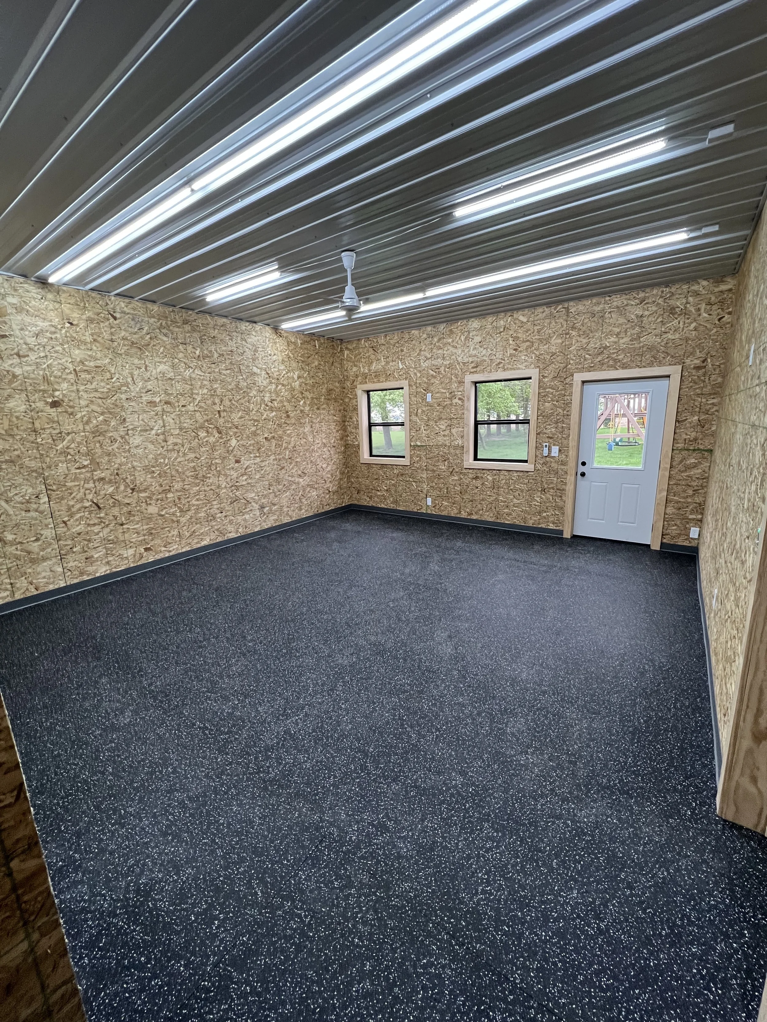 Empty room with plywood walls, two windows, a door, a ceiling fan, and a black speckled rubber floor.