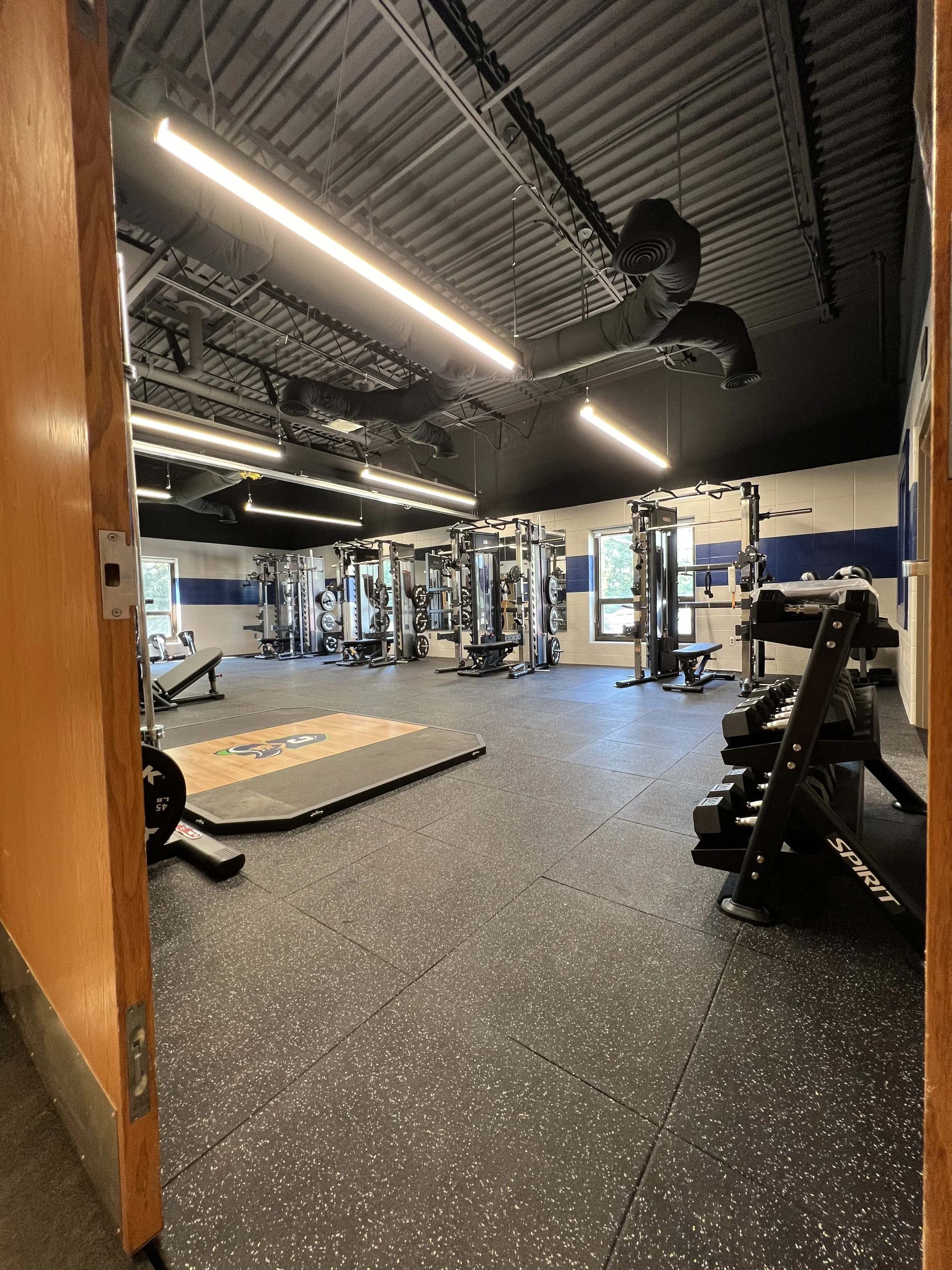 Empty gym room with exercise equipment including weight racks, benches, and a lifting platform, with windows letting in natural light and modern ceiling lights.