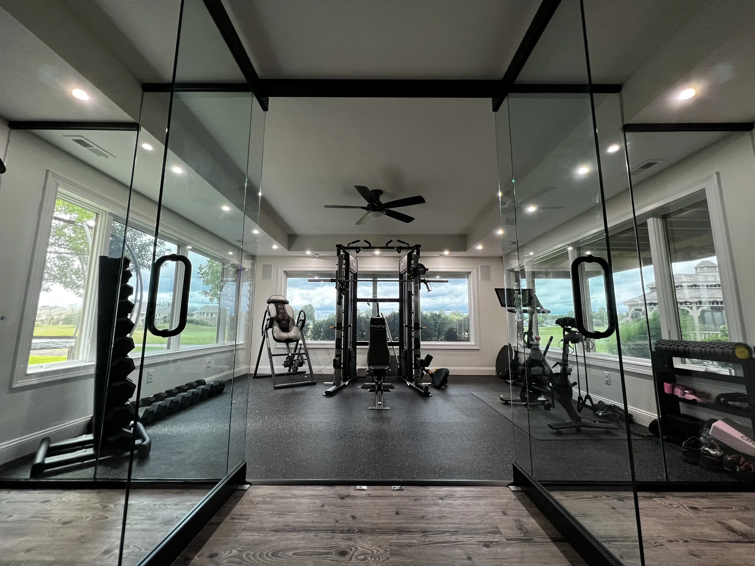 Home gym with various exercise equipment including a weight rack, stationary bike, and weight bench, viewed through glass doors with large windows showing outdoor scenery.