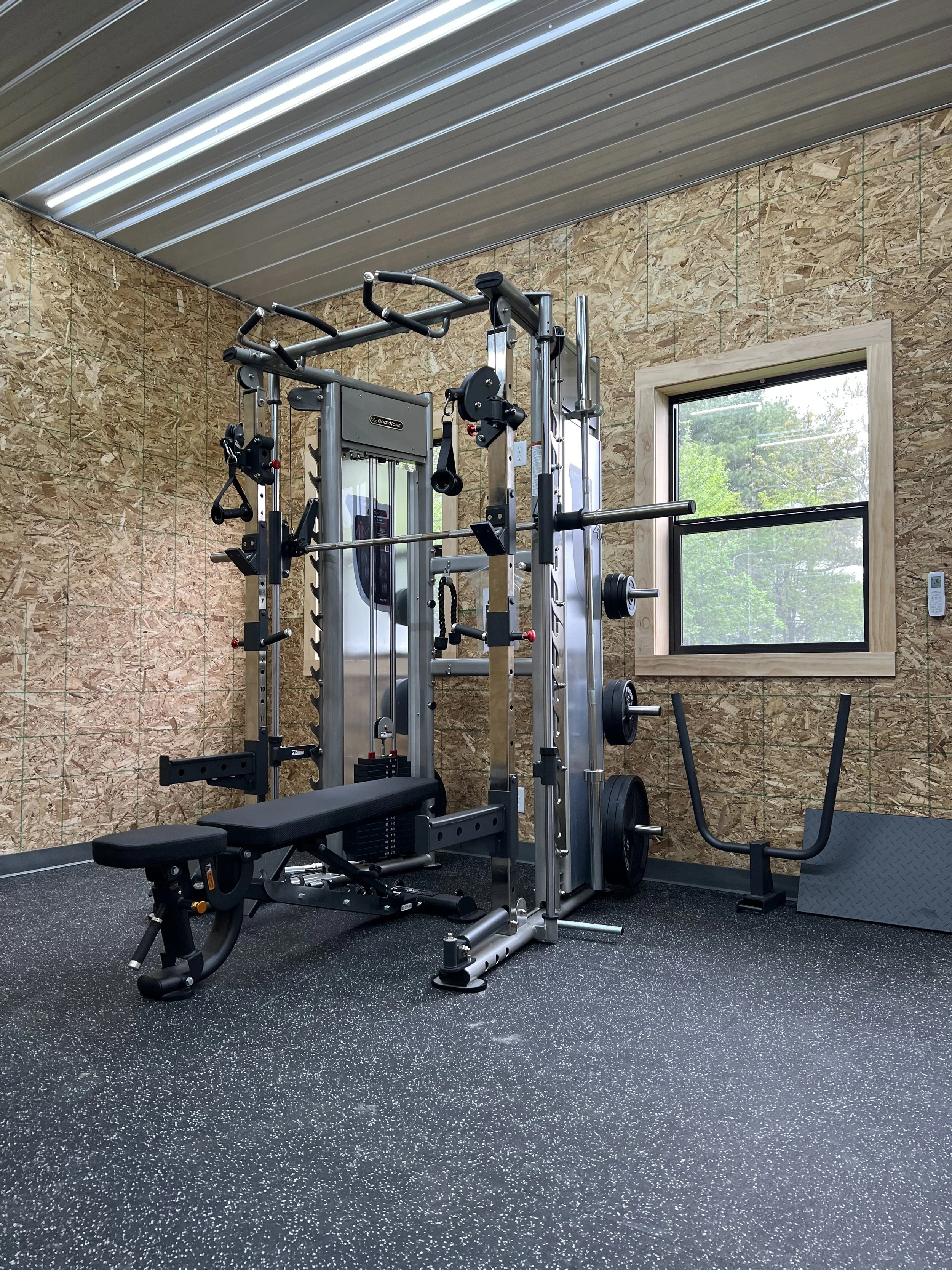 Empty home gym with multi-purpose workout equipment near a window, with black rubber flooring and wooden wall panels.