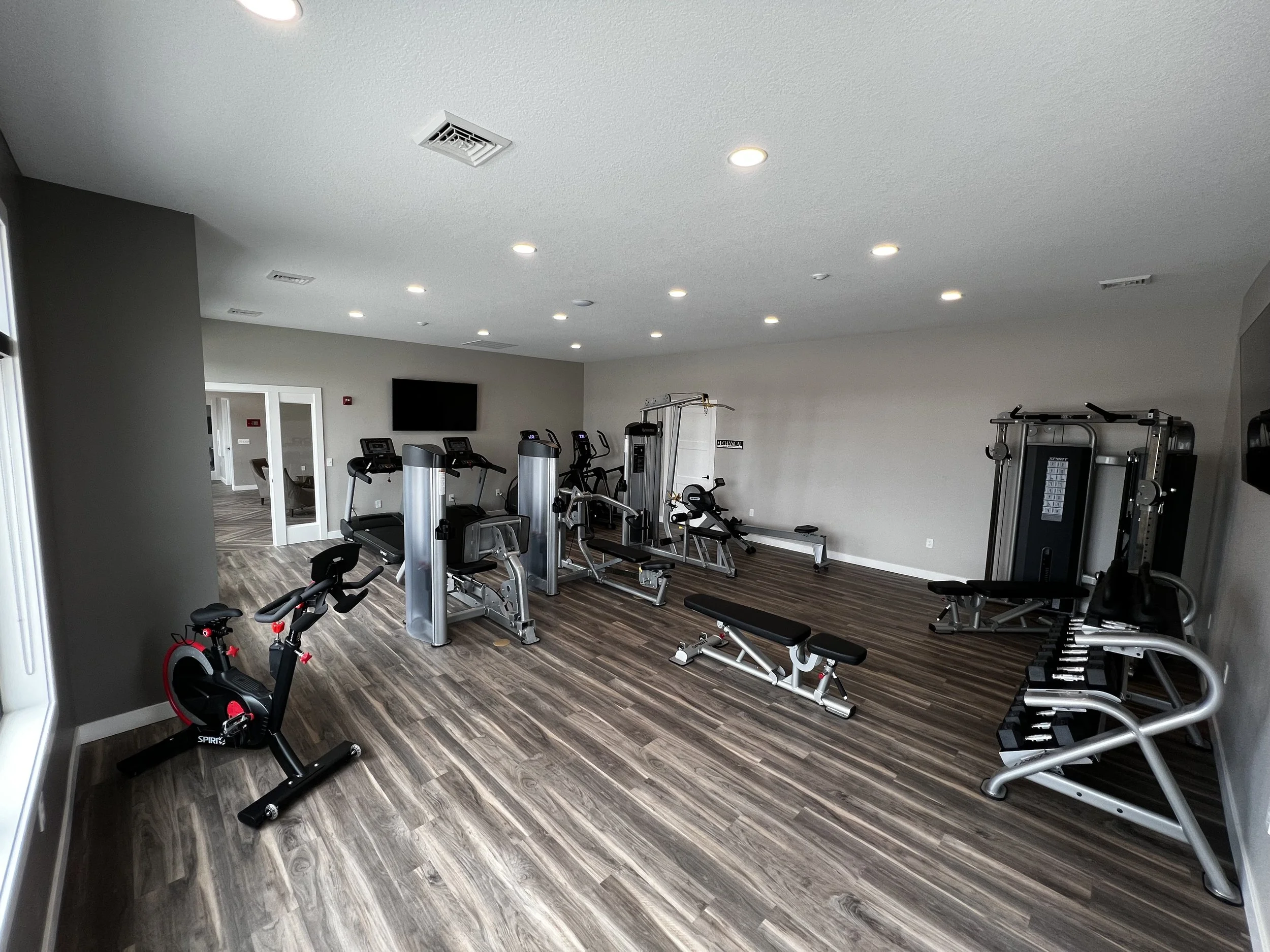 Empty gym room with various exercise equipment including a stationary bike, treadmills, weight machines, a workout bench, and a television on the wall.