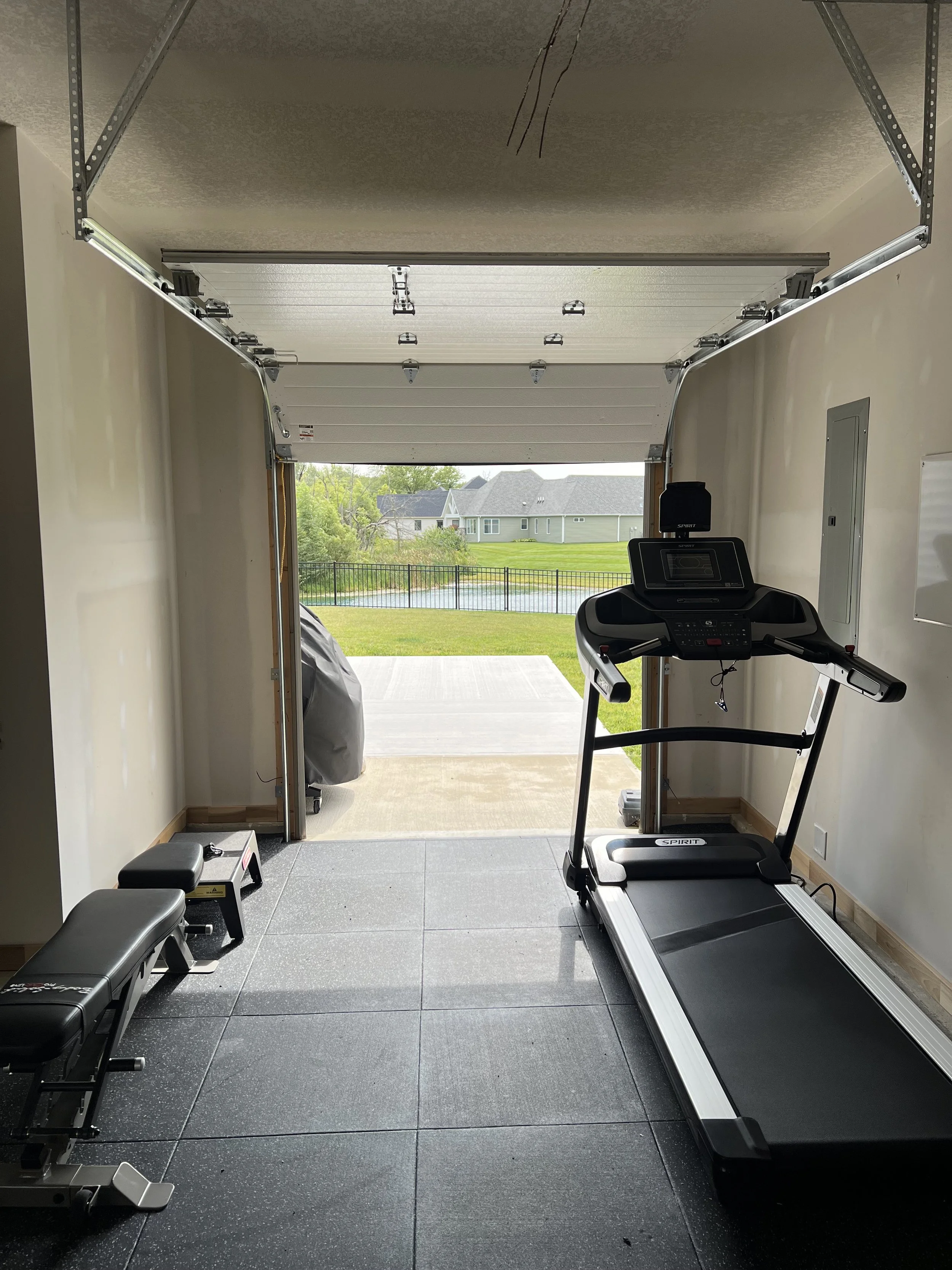 Home gym with treadmill, bench, and weights, inside a garage overlooking a backyard with a pond and houses.