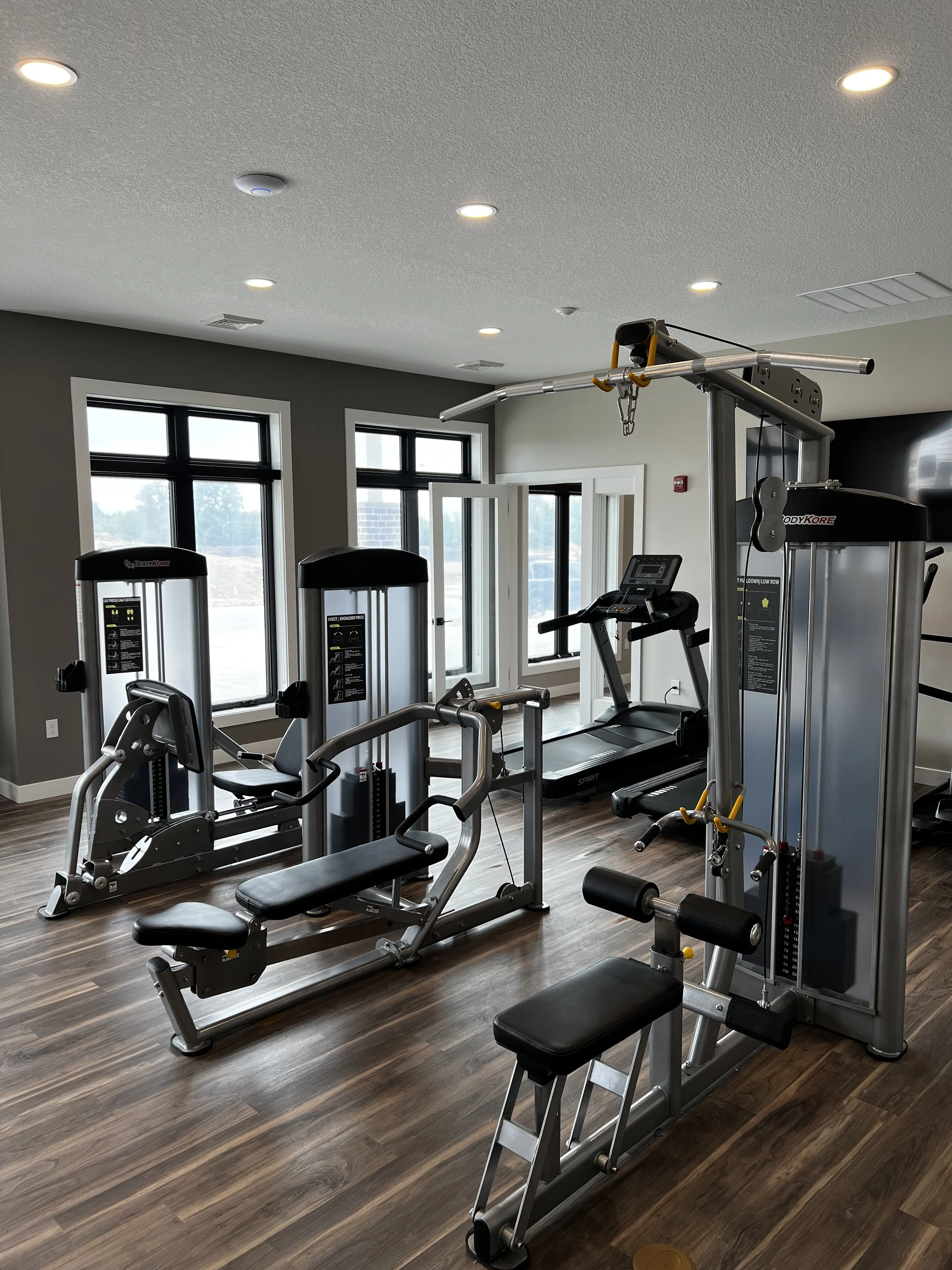 Empty gym with various workout machines, including a treadmill, a seated rowing machine, and weight training equipment, in a room with wooden floors and large windows.