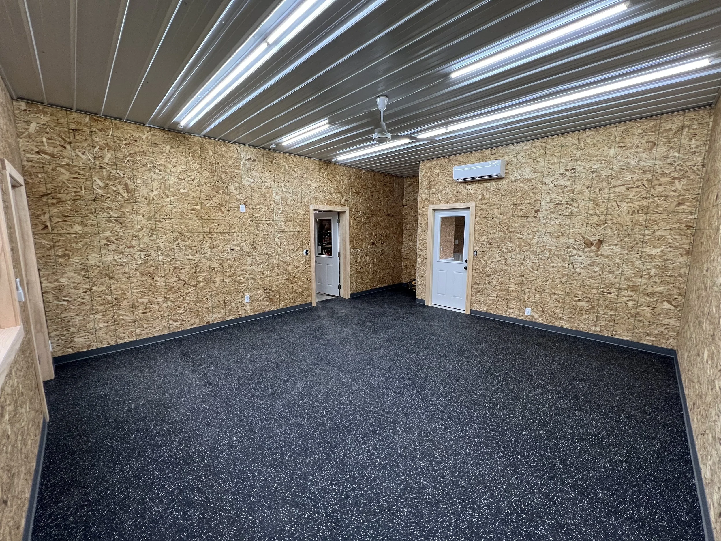 Empty room with unfinished plywood walls, black speckled carpet, metal ceiling, ceiling fan, air conditioning unit, and two white doors.