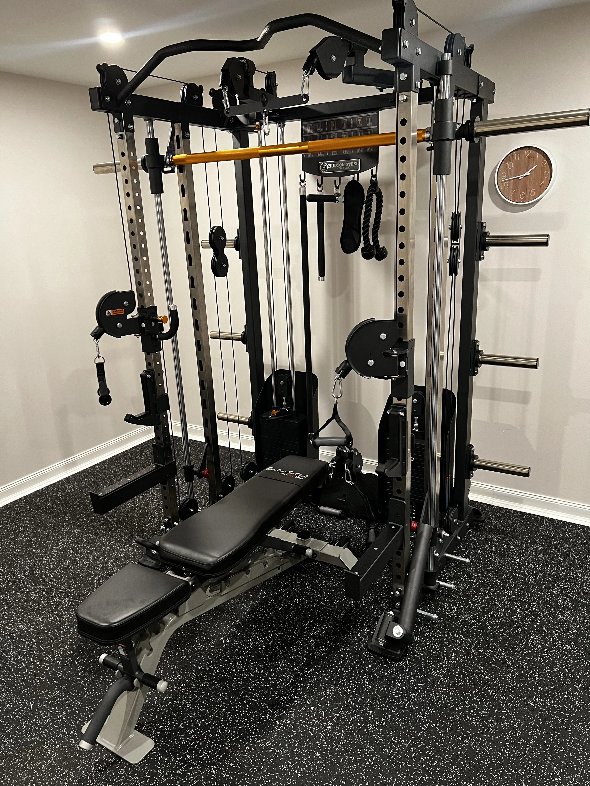A multi-purpose home gym with various workout equipment including a bench, barbells, pulleys, and resistance bands, set on black rubber flooring in a room with white walls and a wall clock.