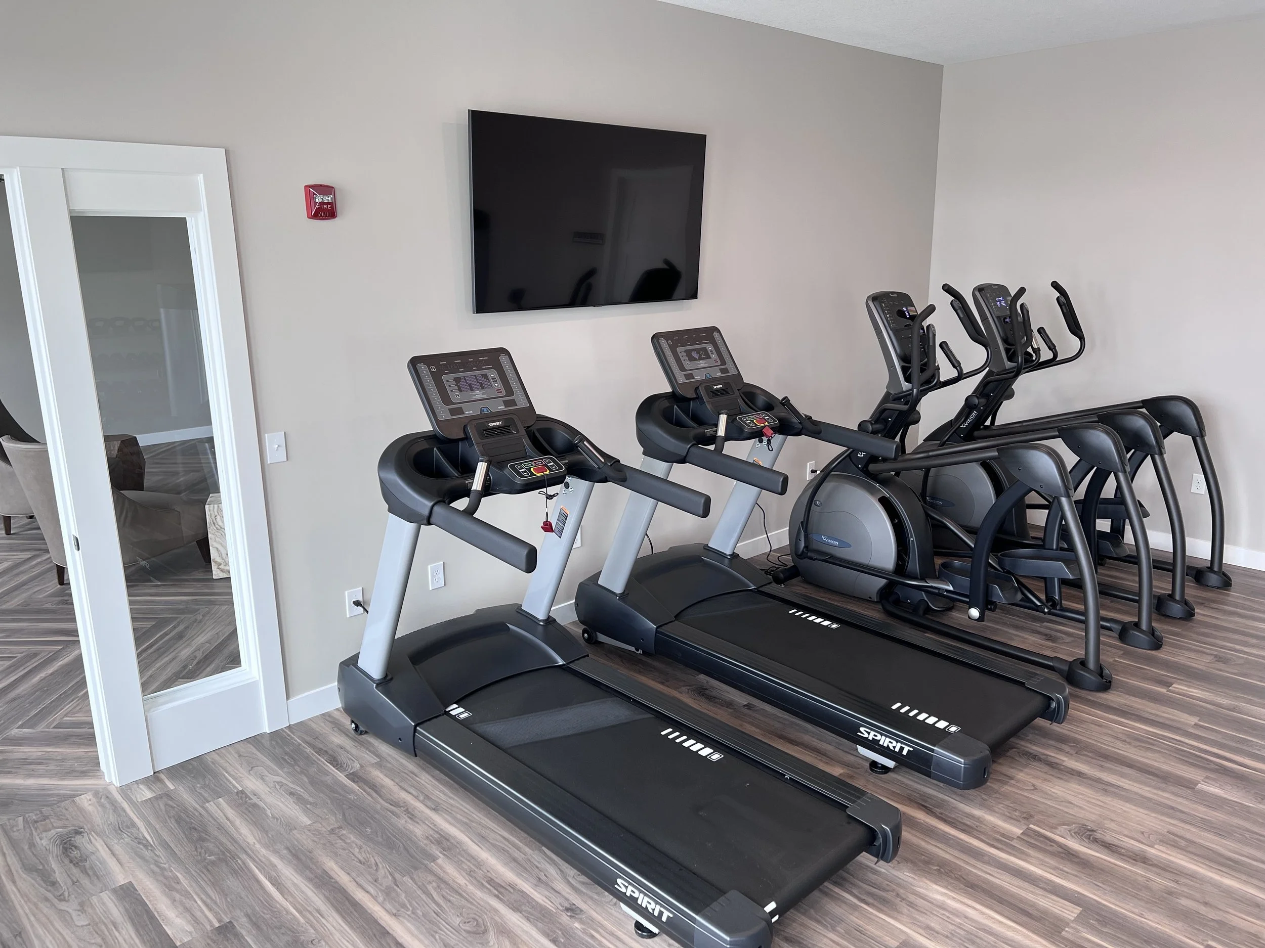 Fitness room with two treadmills and three elliptical machines facing a wall-mounted TV, with a glass door leading to another room.