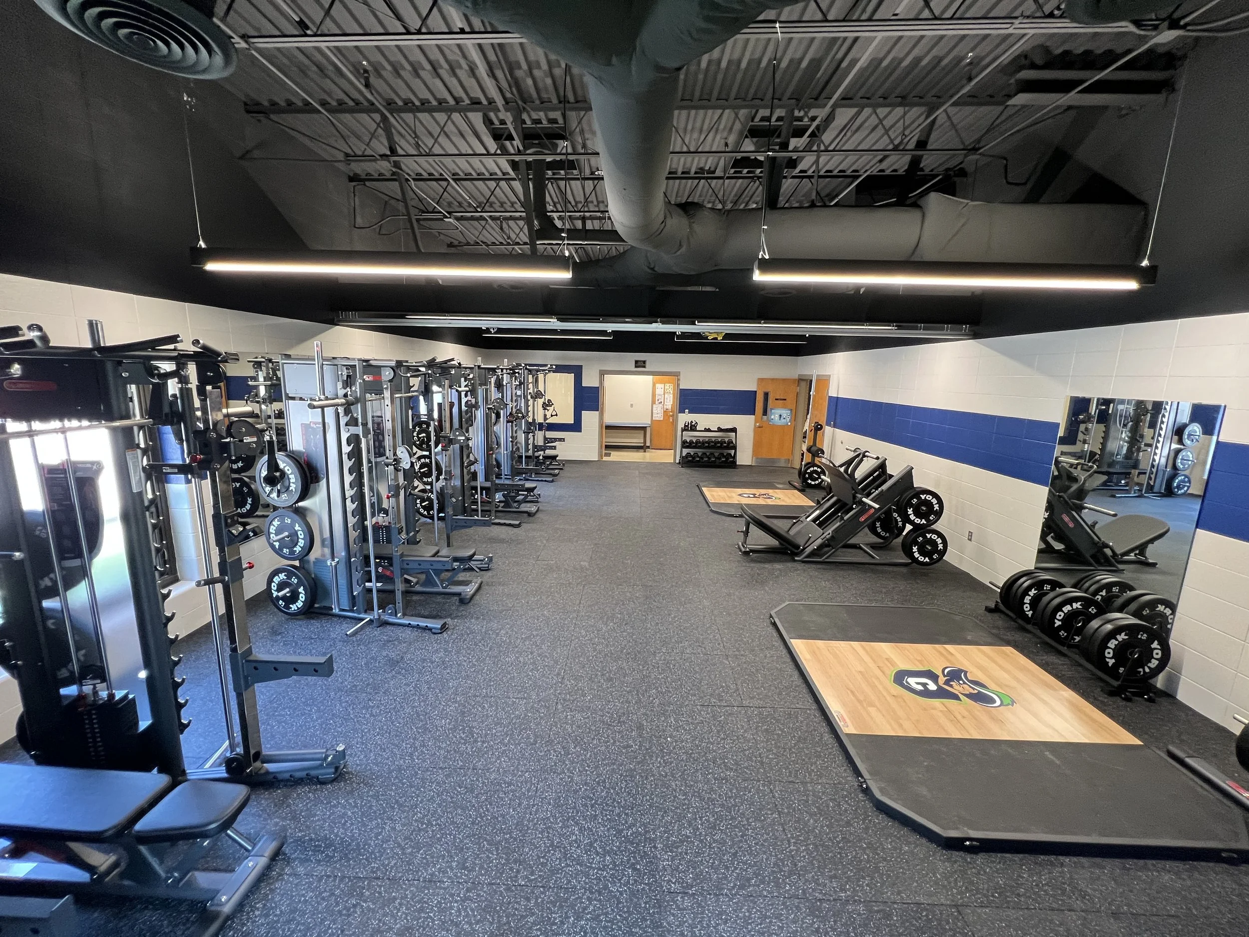 Empty gym with weightlifting equipment, benches, mirrors, and a black ceiling with exposed ductwork.